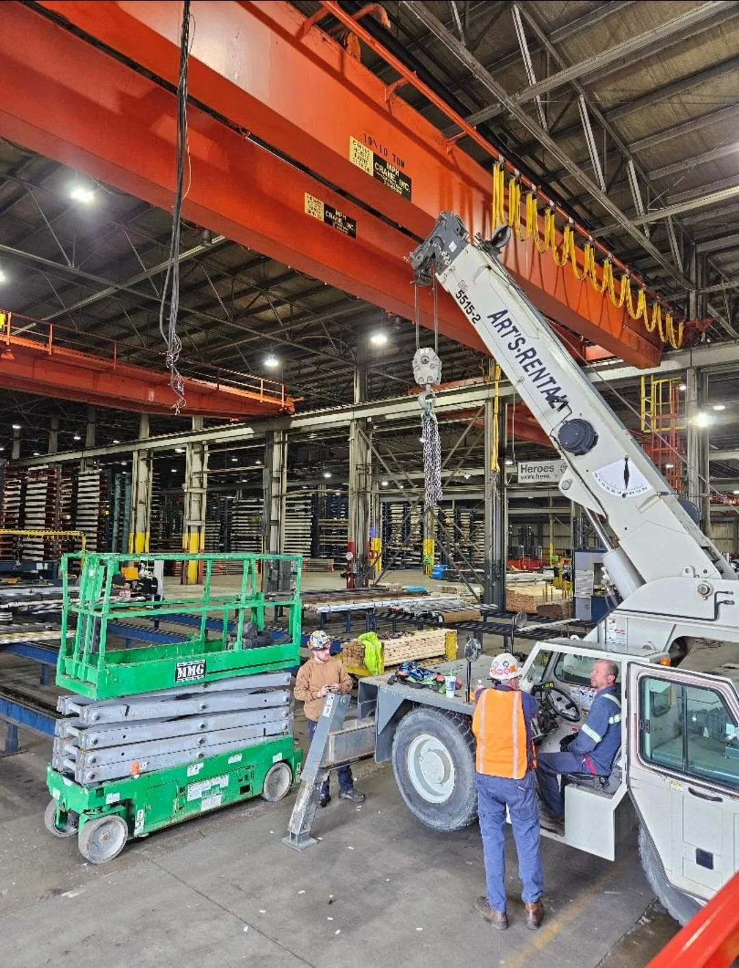 Industrial warehouse with crane lifting materials, workers in safety gear, and various equipment and supplies.