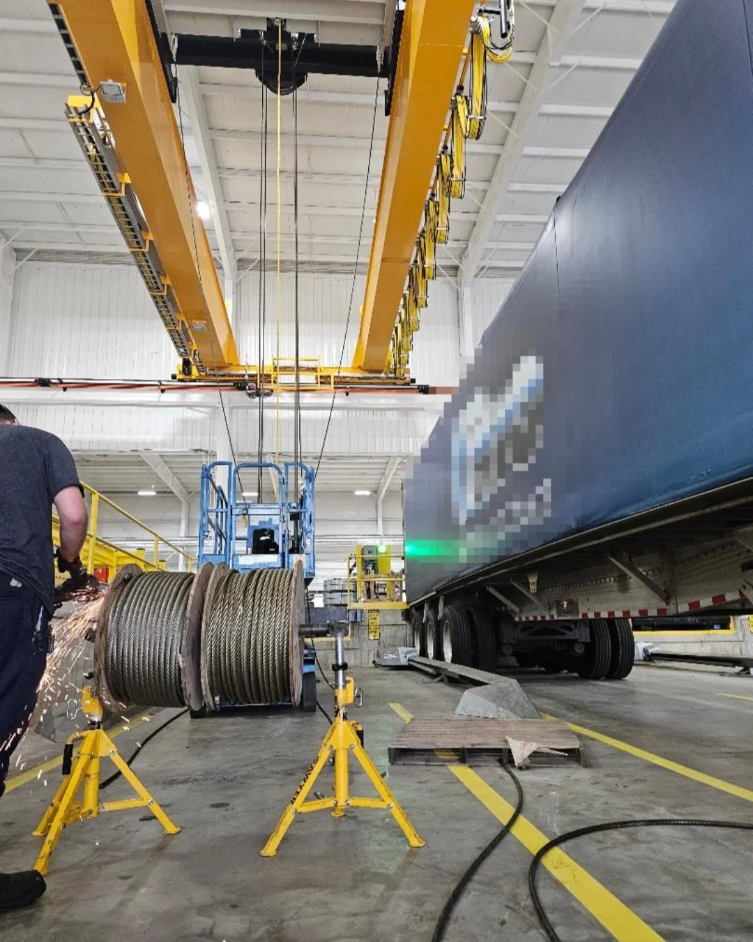 Workers use welding tools in an industrial warehouse with yellow overhead crane equipment and large coiled wires, as a large vehicle with a blurred logo is positioned nearby.