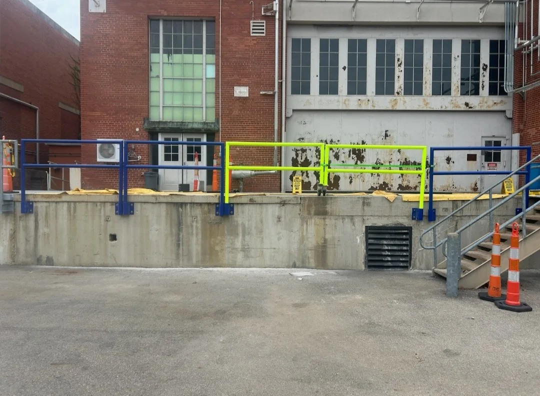View of a concrete loading dock area with blue and green safety railings, orange traffic cones, and industrial stairs on the right. The background features a brick building with large windows, some of which are damaged or covered.