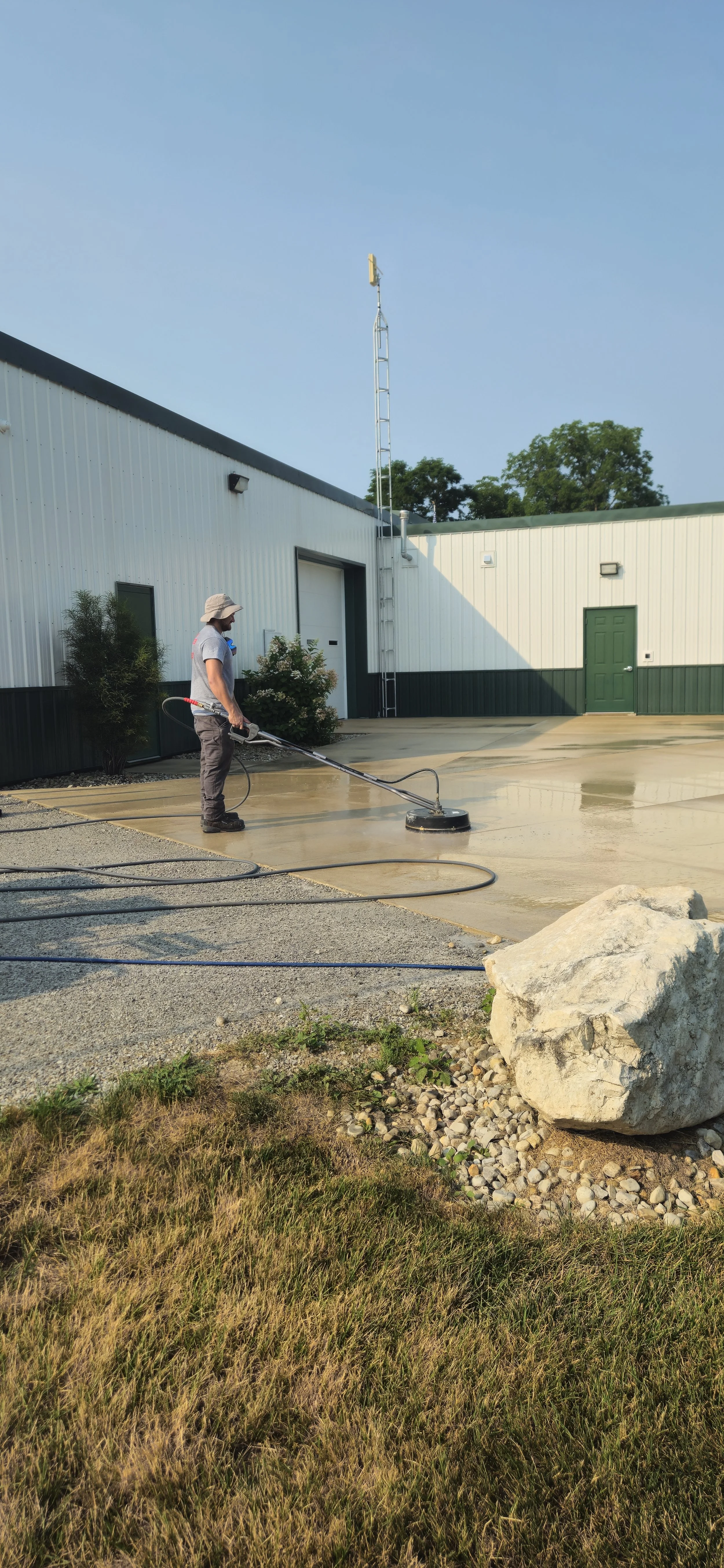 A person is power washing a concrete surface outside a building with a white exterior and green door, under a clear sky.