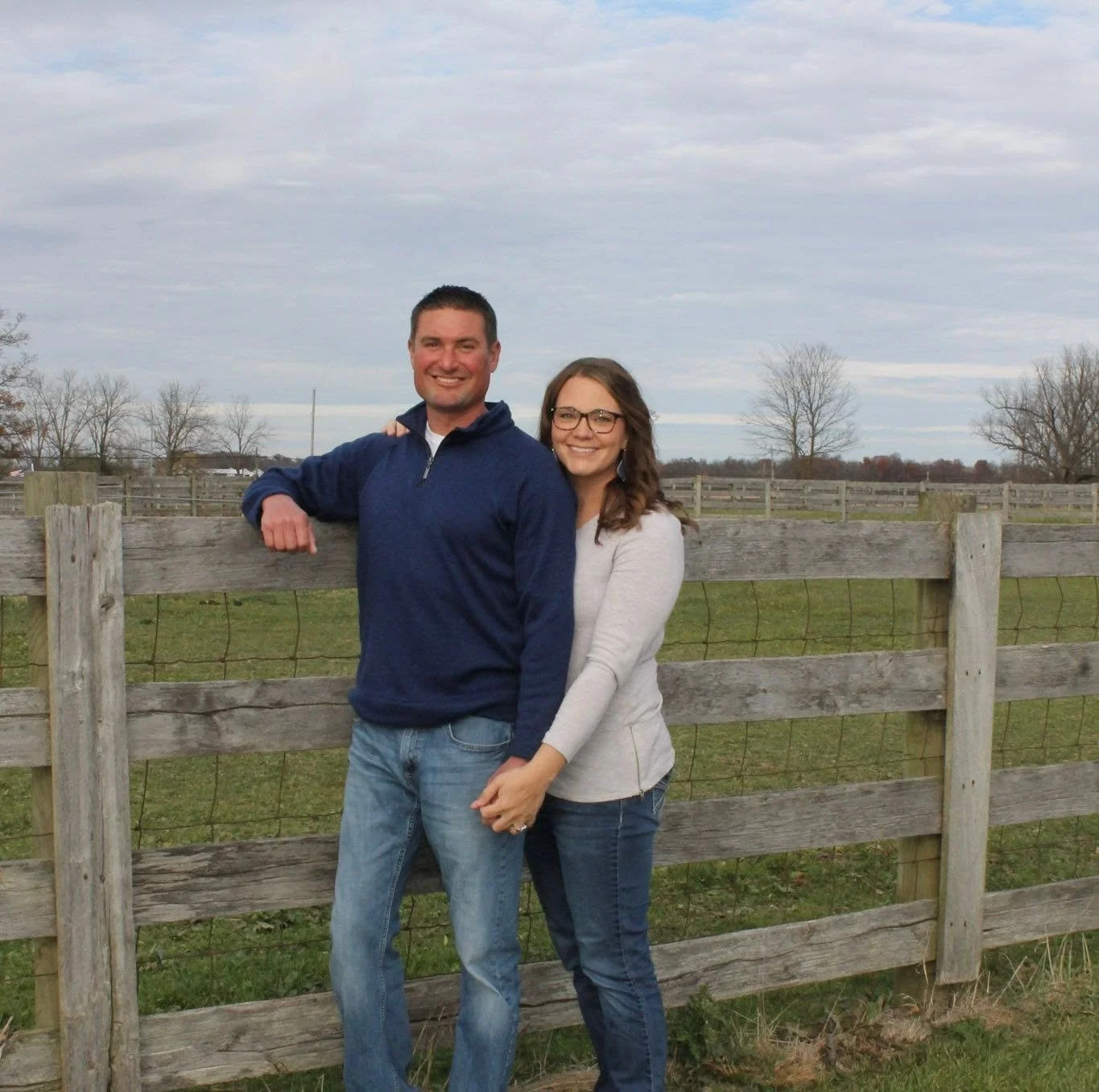 A man and woman standing outdoors in front of a wooden fence, smiling, with a grassy field and trees in the background.