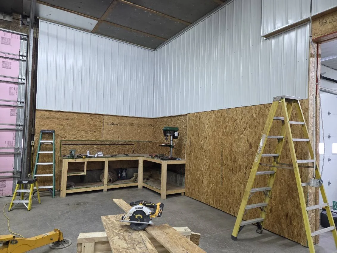 Construction in progress with wooden framing, ladders, power tools, and partially installed wall panels in a workshop or garage.