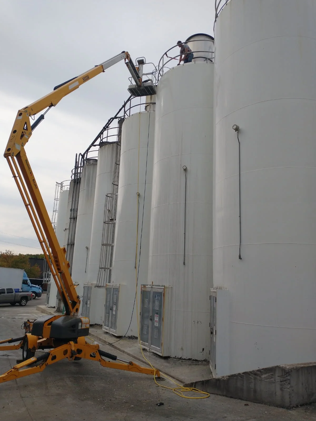Workers on a lift working on large industrial storage tanks outdoors with utility boxes mounted at the base.