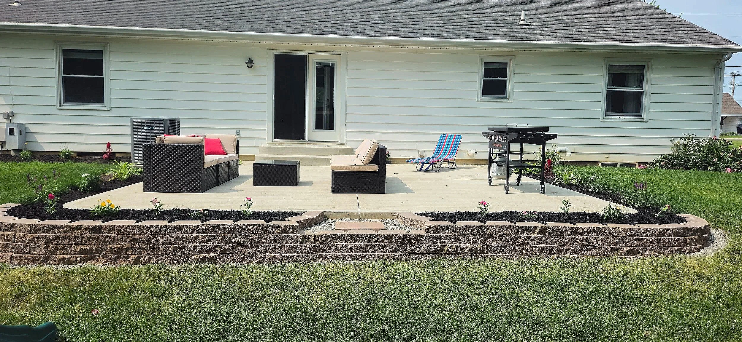 Backyard patio with outdoor furniture, a grill, and garden beds with flowers in front of a house with white siding and three windows.