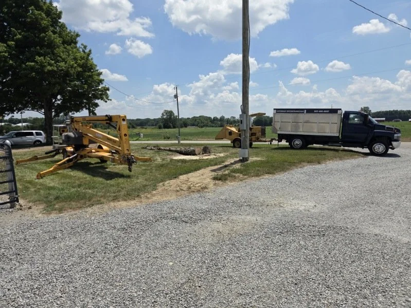 A rural construction site with a large hole in the ground, construction machinery, a utility pole, a black truck, and a field in the background under a partly cloudy sky.