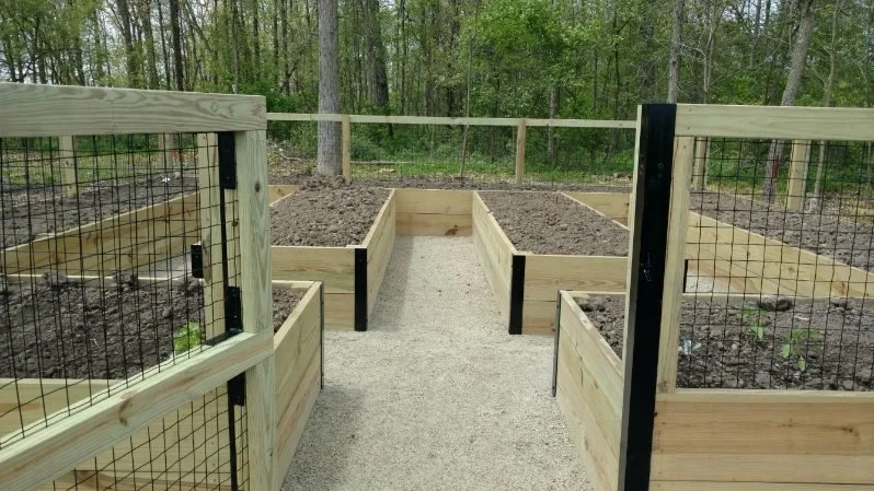 Wooden garden beds with soil, enclosed by a wooden and wire fence, in a wooded outdoor setting.