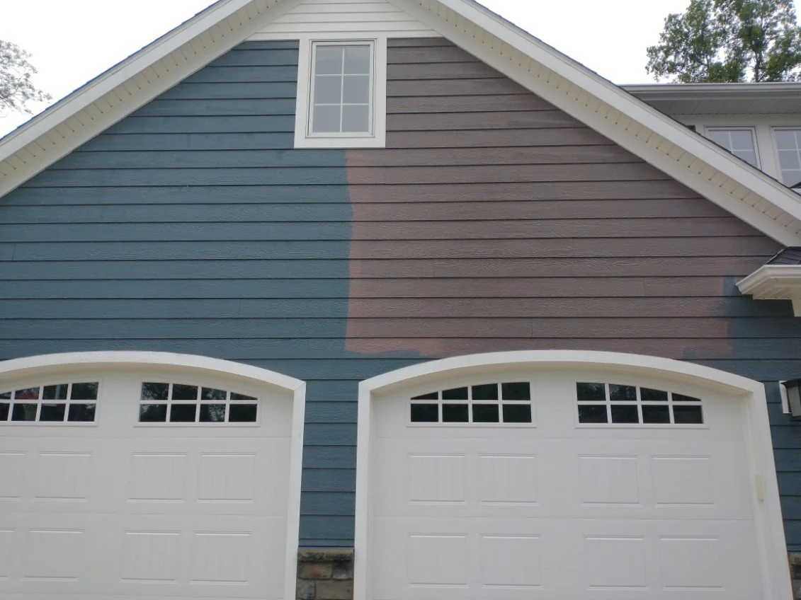 Part of a house showing blue and brown painted siding with two white garage doors below a small window.