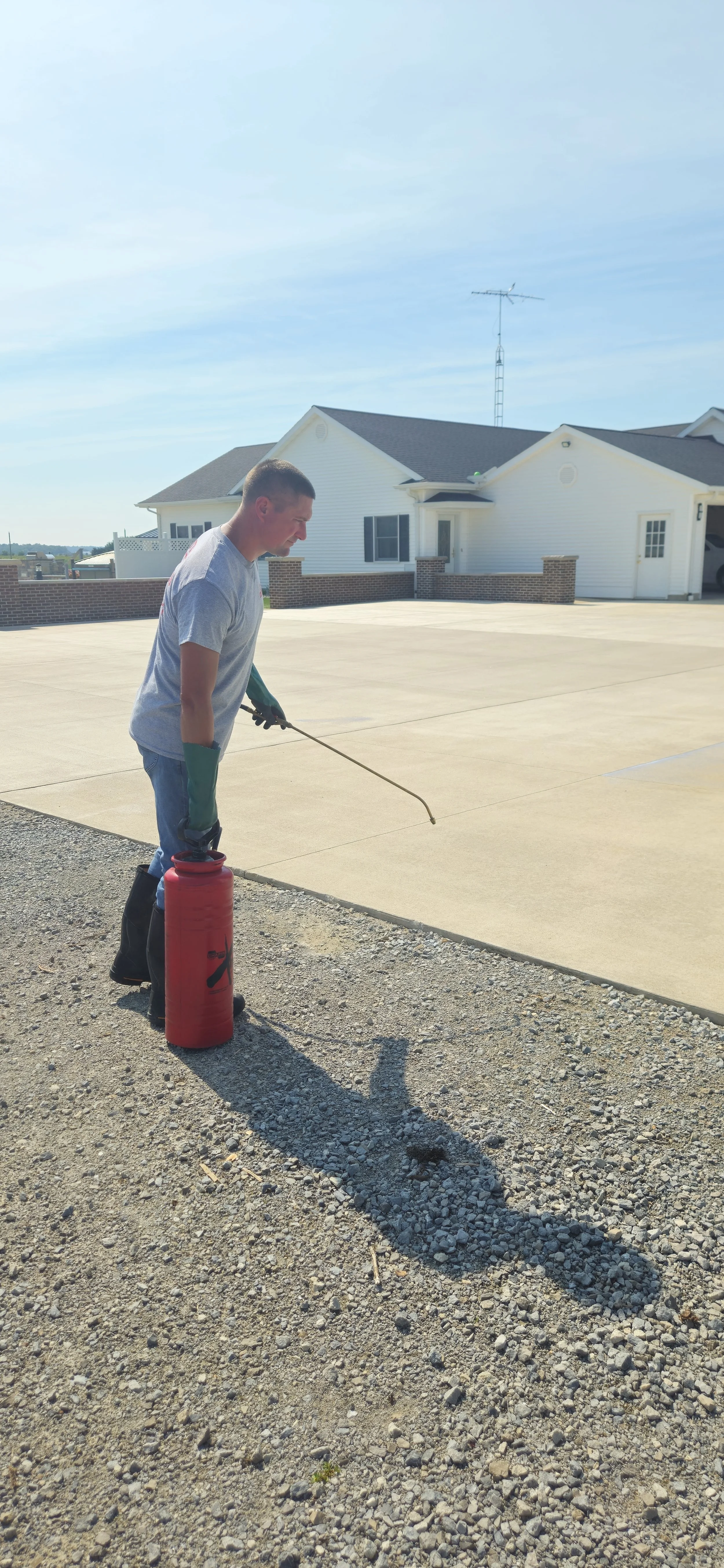 A man wearing gloves and rubber boots is spray painting a concrete driveway with a spray tool connected to a red tank, in front of a white house with a black roof.