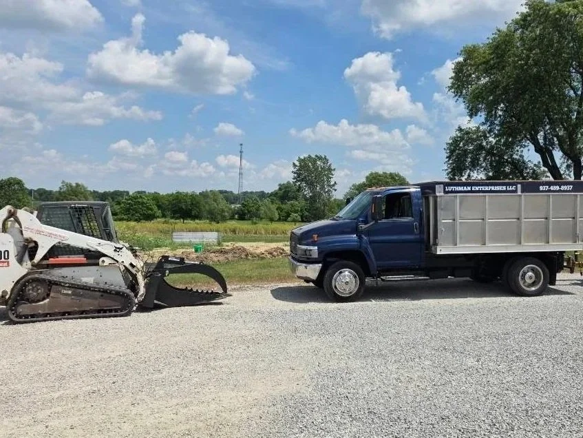 A blue dump truck parked on a gravel road facing a small tracked construction vehicle against a background of green fields, trees, and cloudy sky.