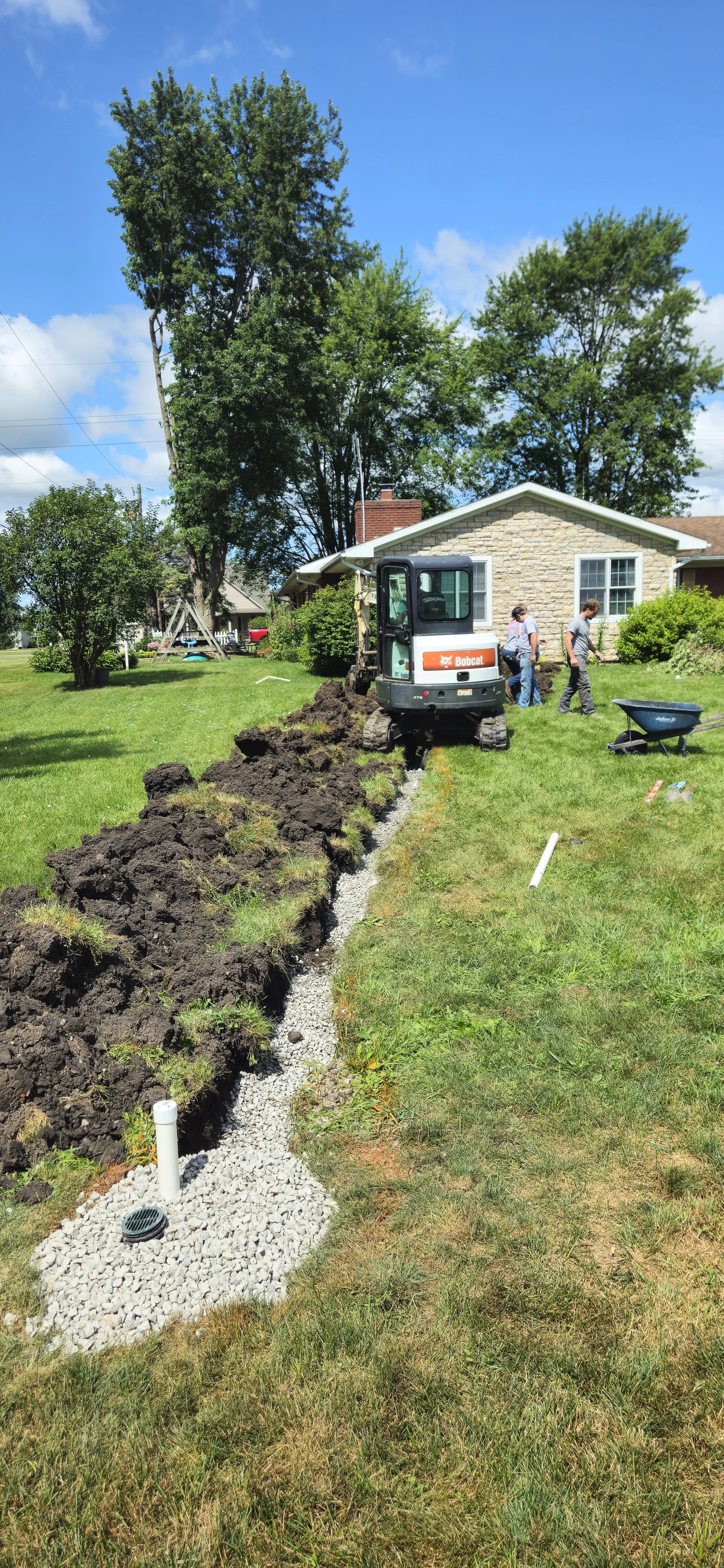 Workers installing a gravel drainage trench alongside a house lawn with a small excavator and dirt piled up.