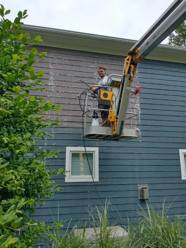 Man on a lift working on the exterior wall of a house with blue siding, while a tree and some grass are in the foreground.