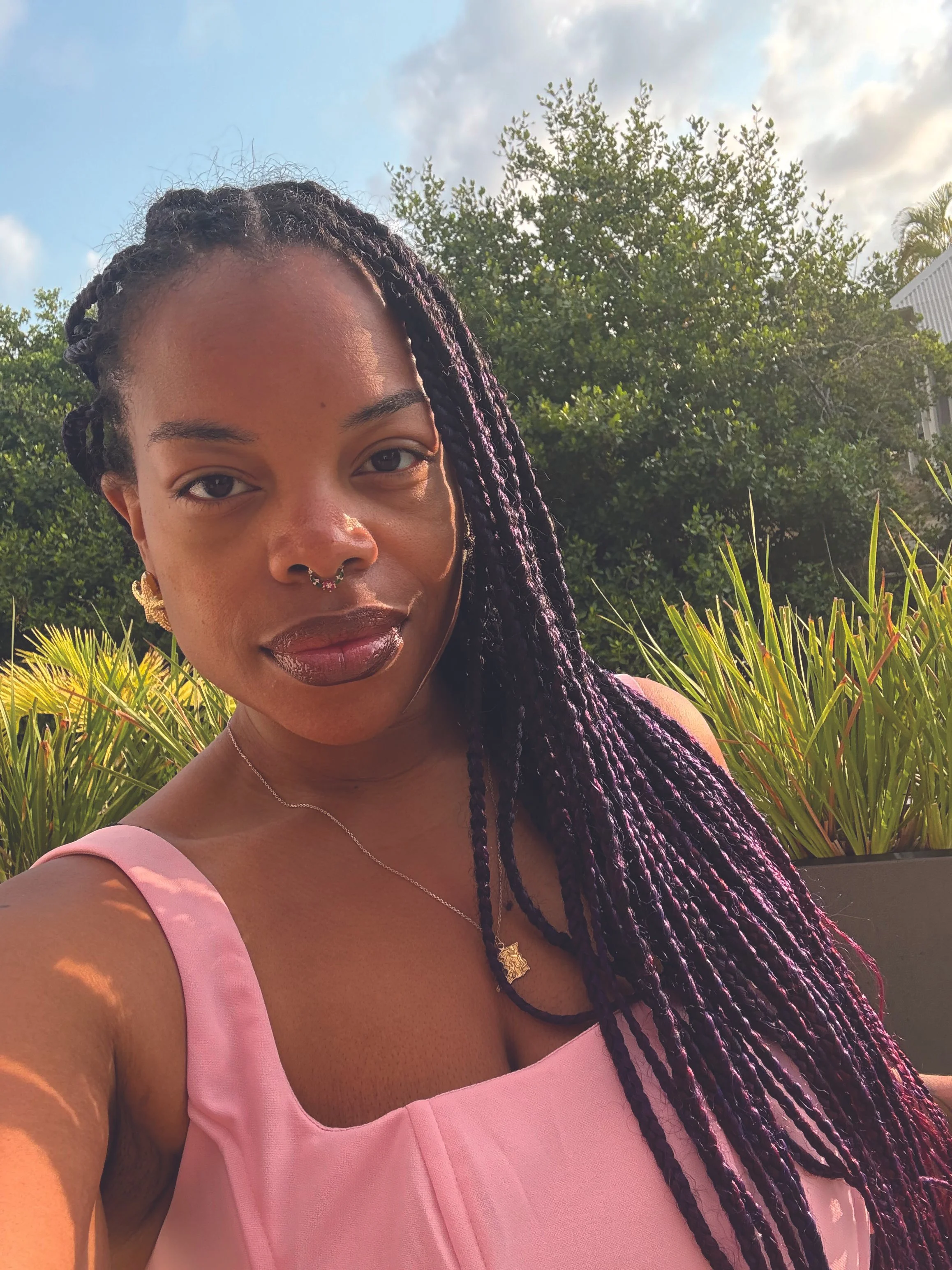 Close-up of a woman with braided purple-tinted hair, jewelry including earrings, a septum ring, a necklace, wearing a pink sleeveless top, outdoors with green plants and trees in the background.