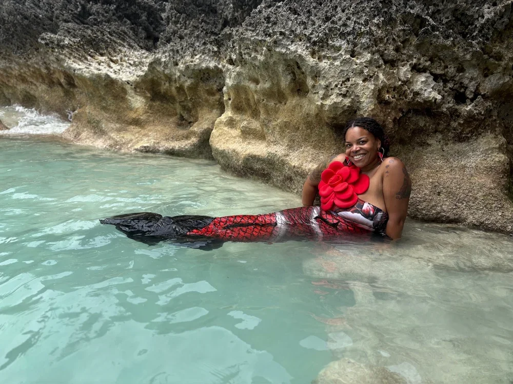 Woman in a mermaid costume sitting in water next to a rocky cave wall, smiling.