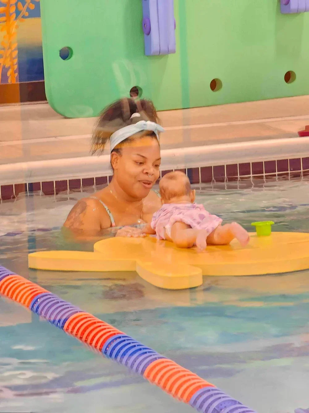 A woman and a baby in a swimming pool. The woman is holding the baby on a yellow foam floatation device, and they are near the edge of the pool with a colorful pool lane divider in the foreground.