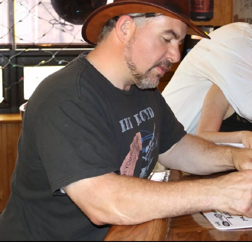 A man wearing a black T-shirt with a graphic design, a wide-brimmed hat, and a beard, is sitting at a wooden table, writing or drawing on a piece of paper. Another person is partially visible next to him, and the setting appears to be indoors with a window and some trees outside in the background.