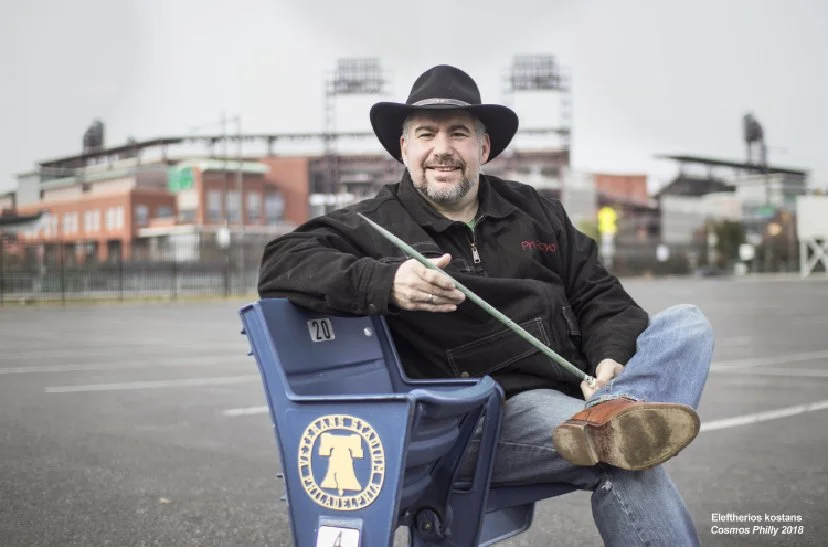 A man sitting in a blue stadium seat on an empty outdoor parking lot, wearing a black hat and jacket, smiling, holding a pointer stick, with a cityscape and sports stadium lights in the background.