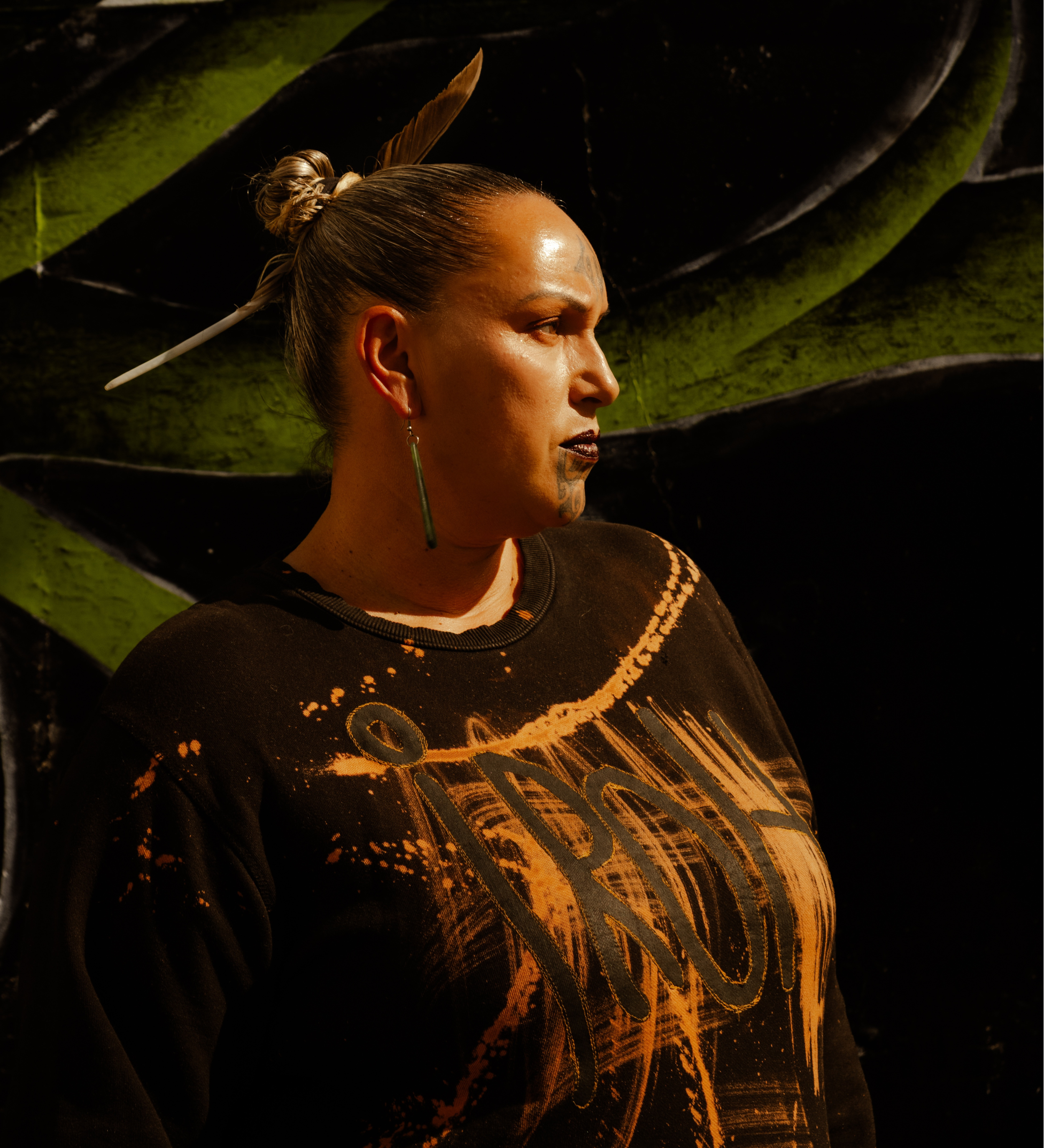 A woman with a painted face and hair in a bun, wearing earrings, stands against a black and green abstract background.