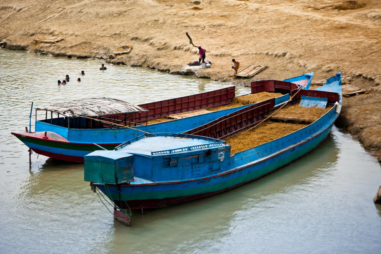 Two large blue boats docked on shallow water near a sandy beach, with people on the beach and in the water, some swimming and others preparing to fish or work.
