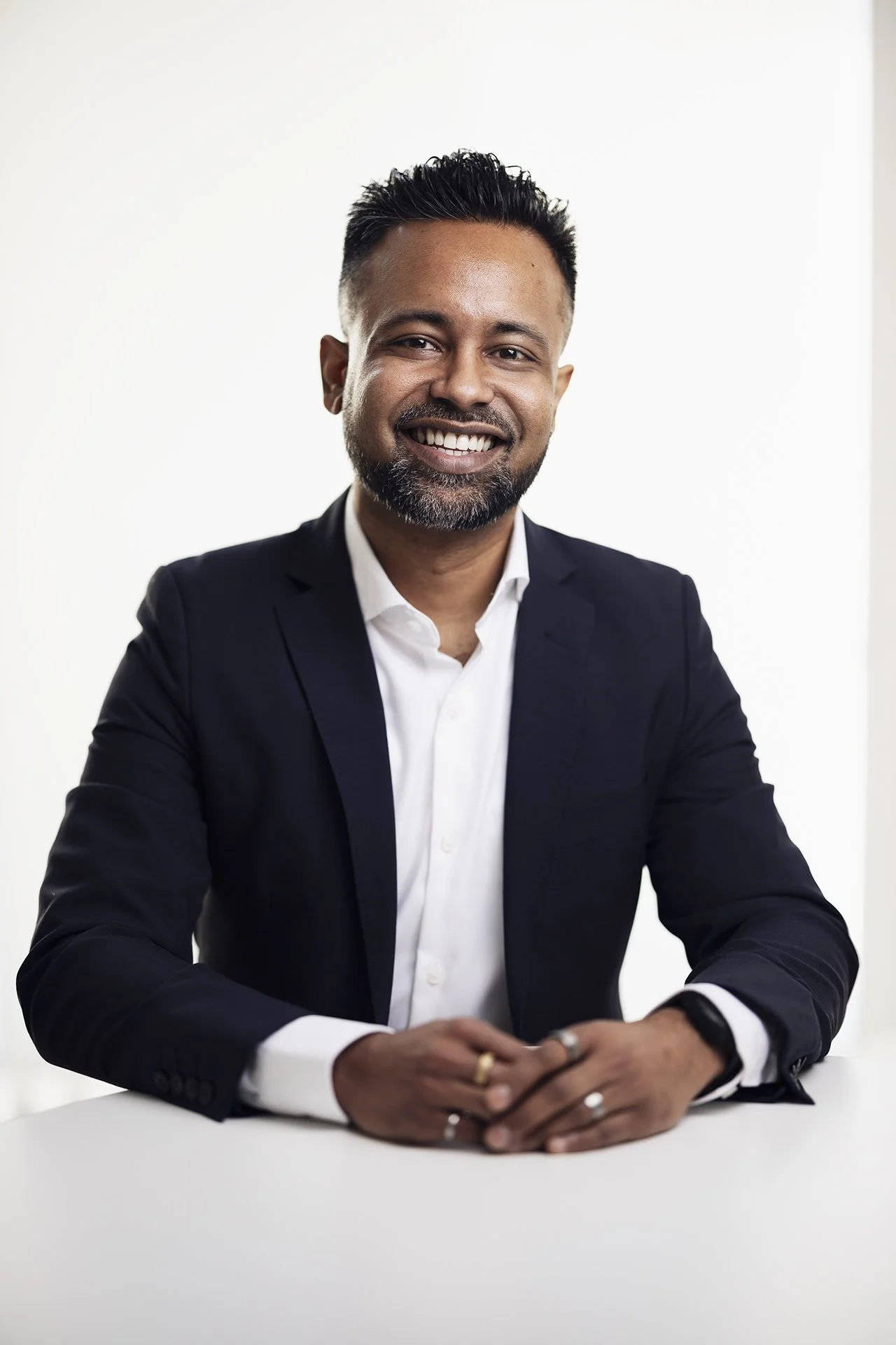 A smiling man in a dark suit with a white shirt, sitting at a white table against a plain white background.