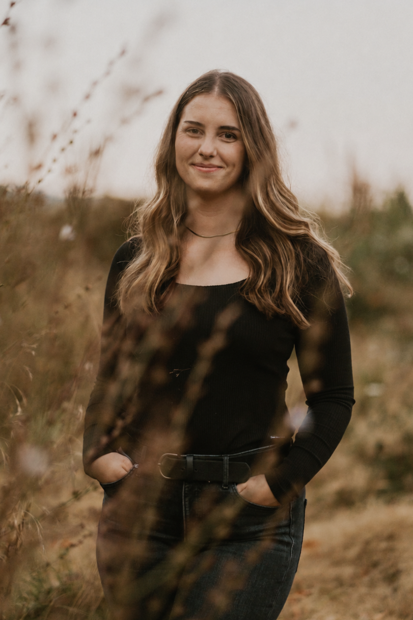 A young woman with long, wavy brown hair standing outdoors in a natural setting during fall, wearing a black long-sleeve top and dark jeans, with her hands in her pockets, smiling gently at the camera.