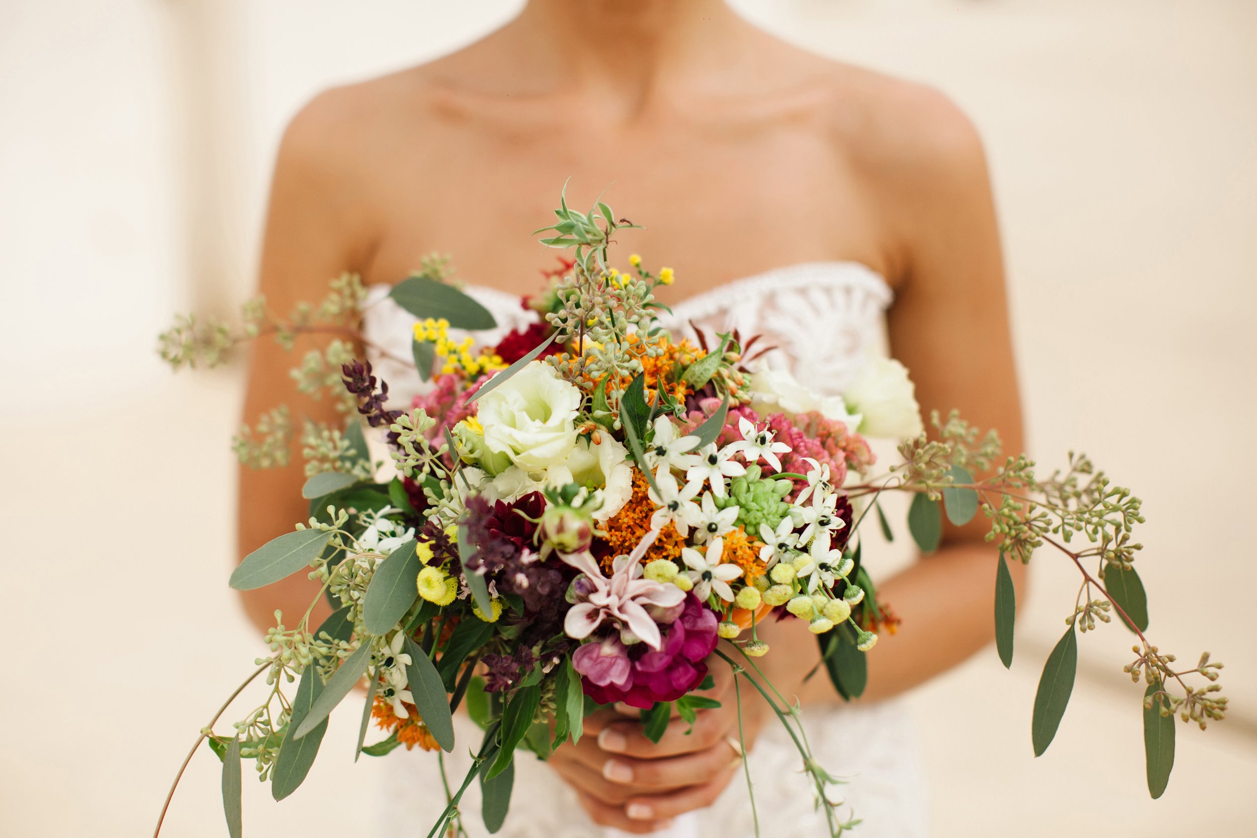 A woman wearing a white strapless dress holding a large, colorful flower bouquet with various flowers and green foliage.