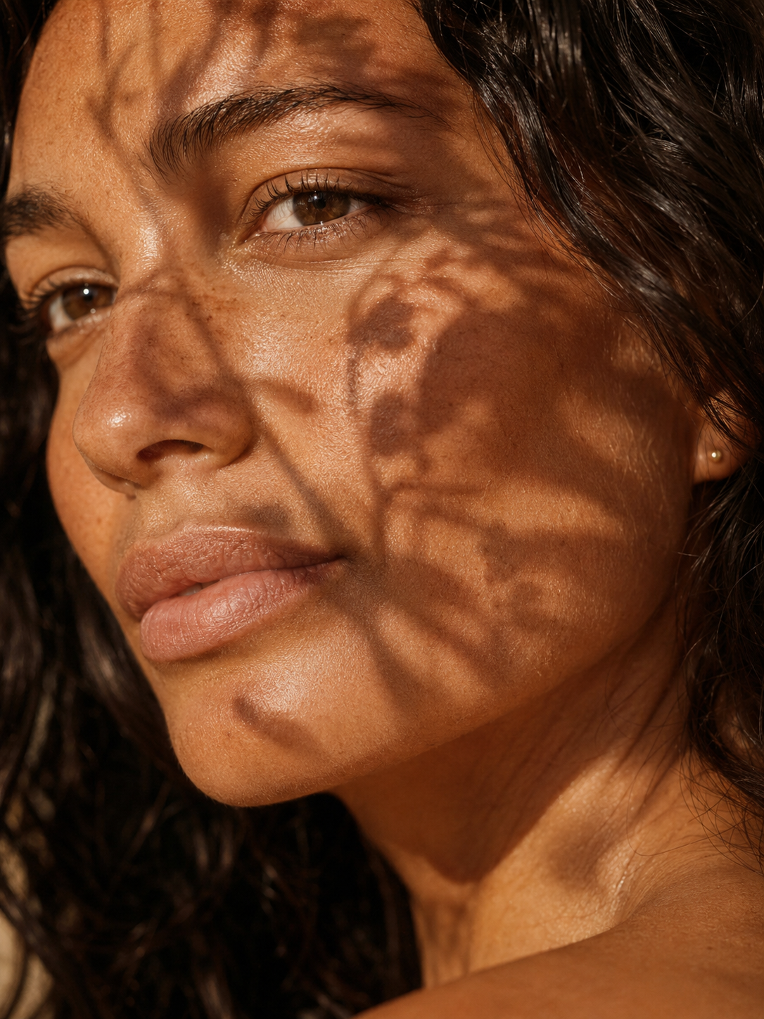 Close-up of a woman's face with sunlight and shadow patterns across her skin, brown eyes, and dark hair.