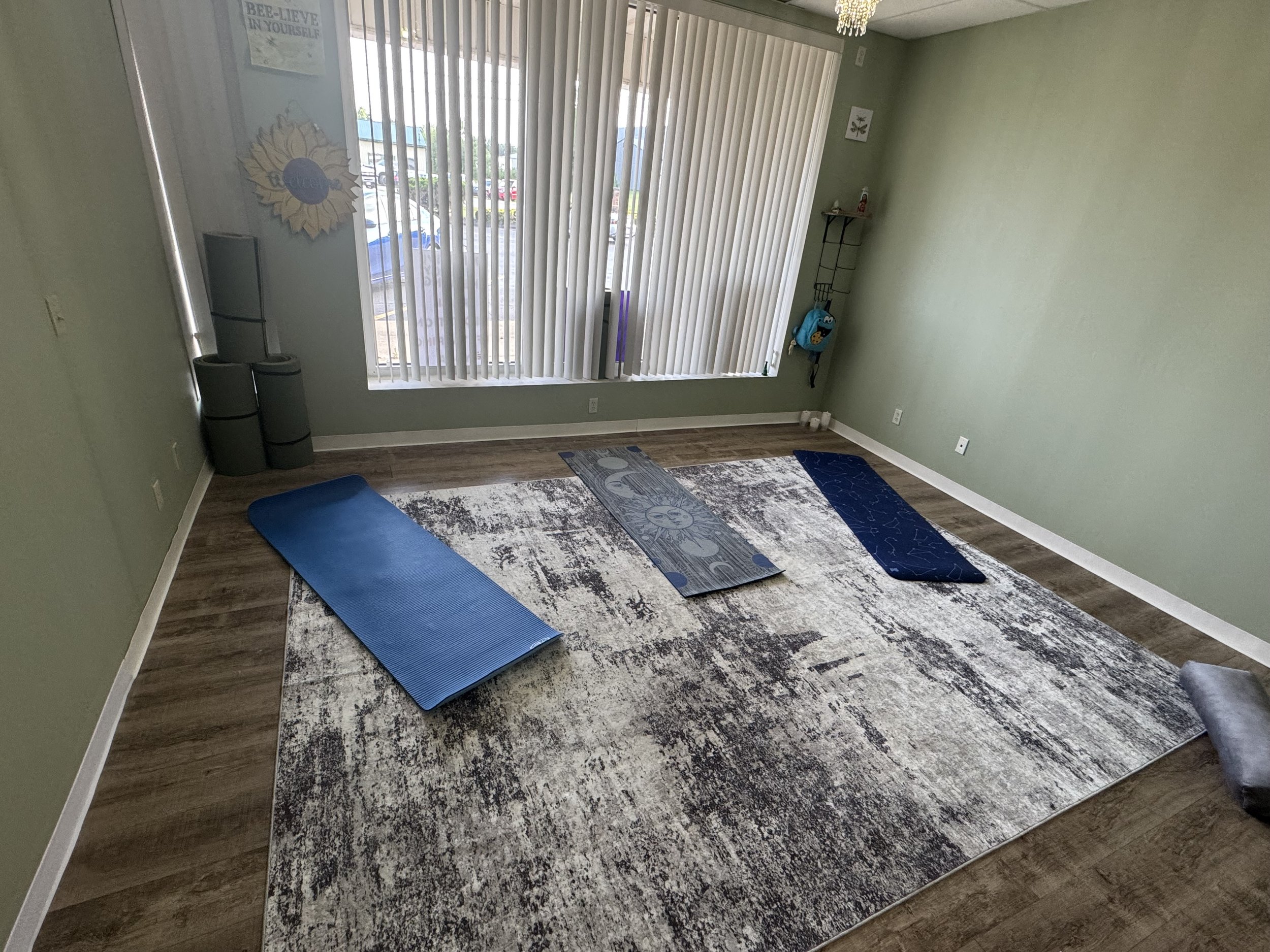 A room with green walls and vertical blinds on large windows that let in natural light. There are yoga mats laid out on a multicolored rug on a wooden floor. A small shelf with decor and a backpack is in the corner, and a poster on the wall reads 'BEE-LIEVE IN YOURSELF.'