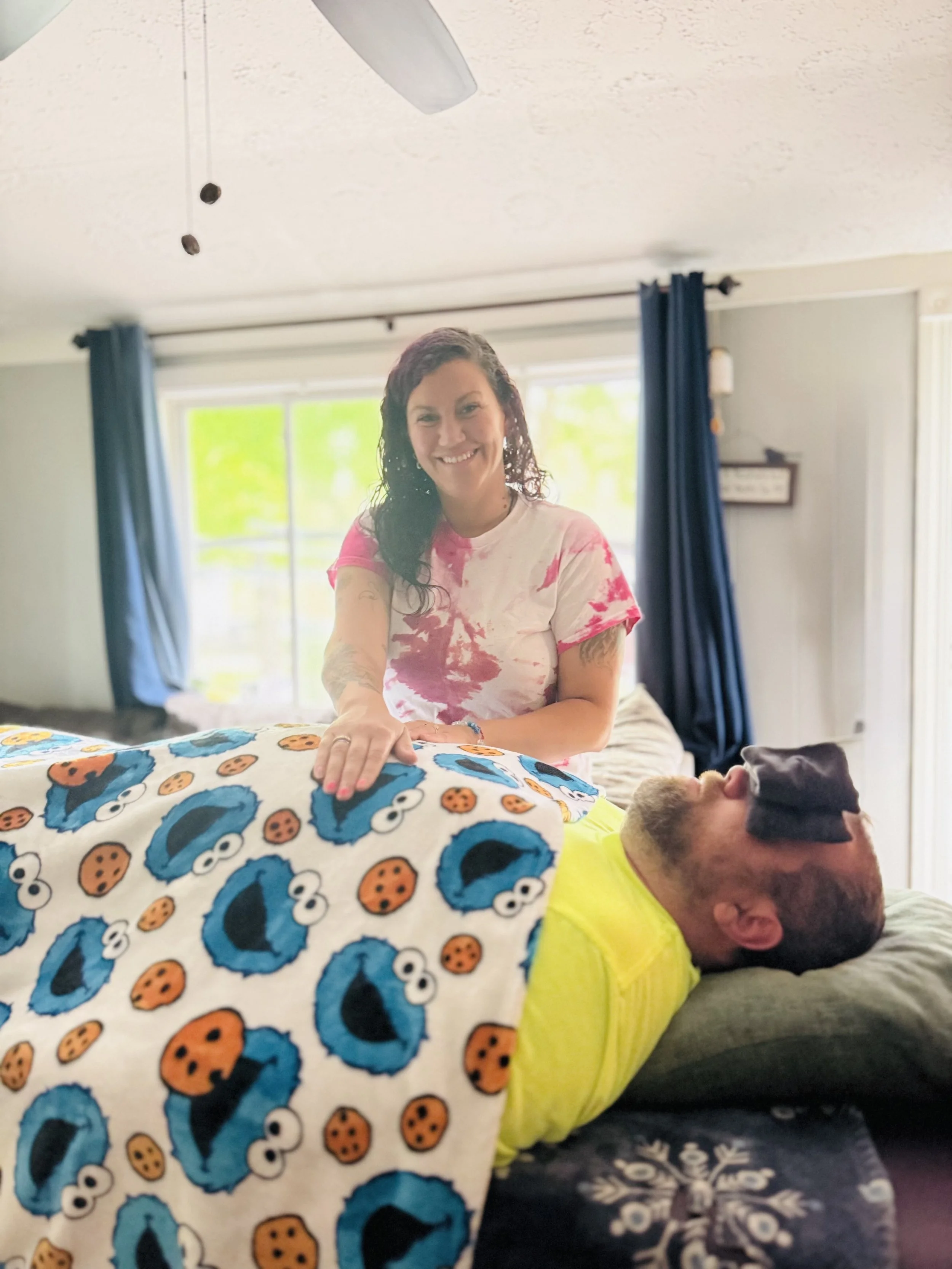 A woman with dark wavy hair, wearing a pink tie-dye shirt, smiling as she helps a man lying on a bed. The man has a beard and is wearing a bright yellow shirt. They are in a bedroom with large windows, blue curtains, and sunlight streaming in.