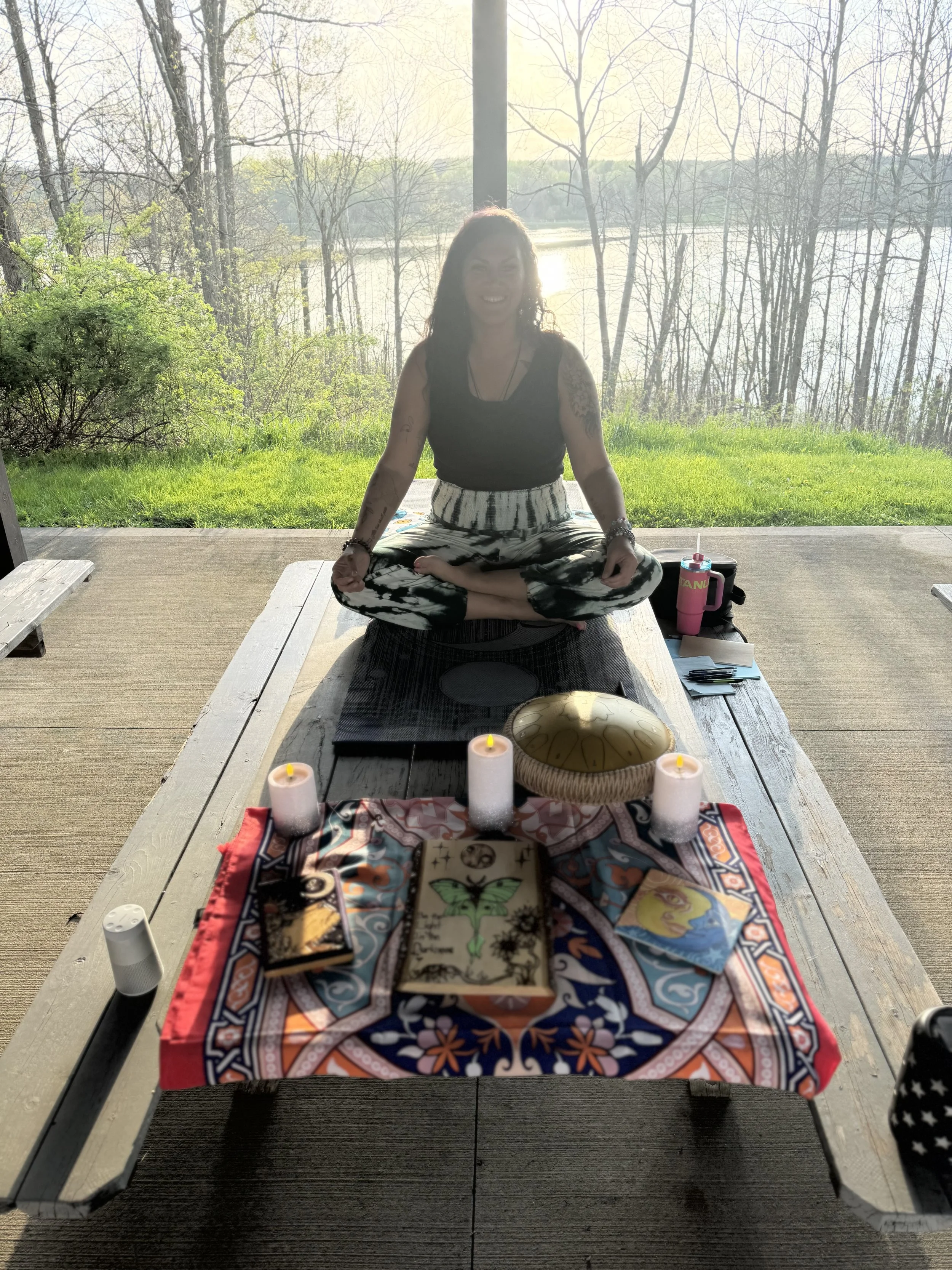 A woman practicing yoga or meditation outdoors on a wooden platform near a body of water, with trees and sunlight in the background. She is sitting cross-legged on a yoga mat, smiling, with candles and tarot cards placed on a decorative cloth in front of her.