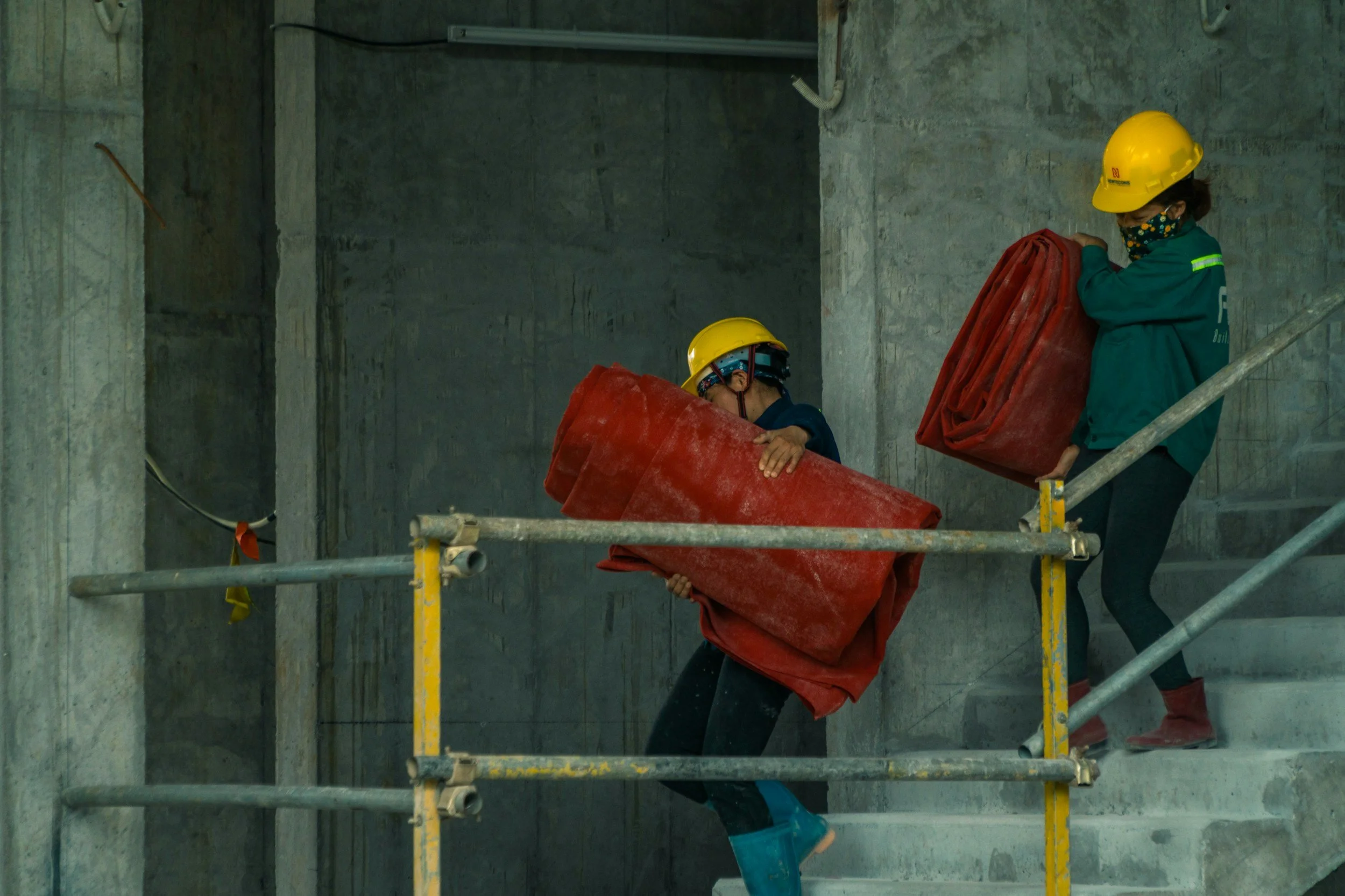 Two construction workers wearing yellow hard hats and masks are handling red safety barriers inside a building under construction. One worker is holding a barrier while the other is placing it on a scaffold.