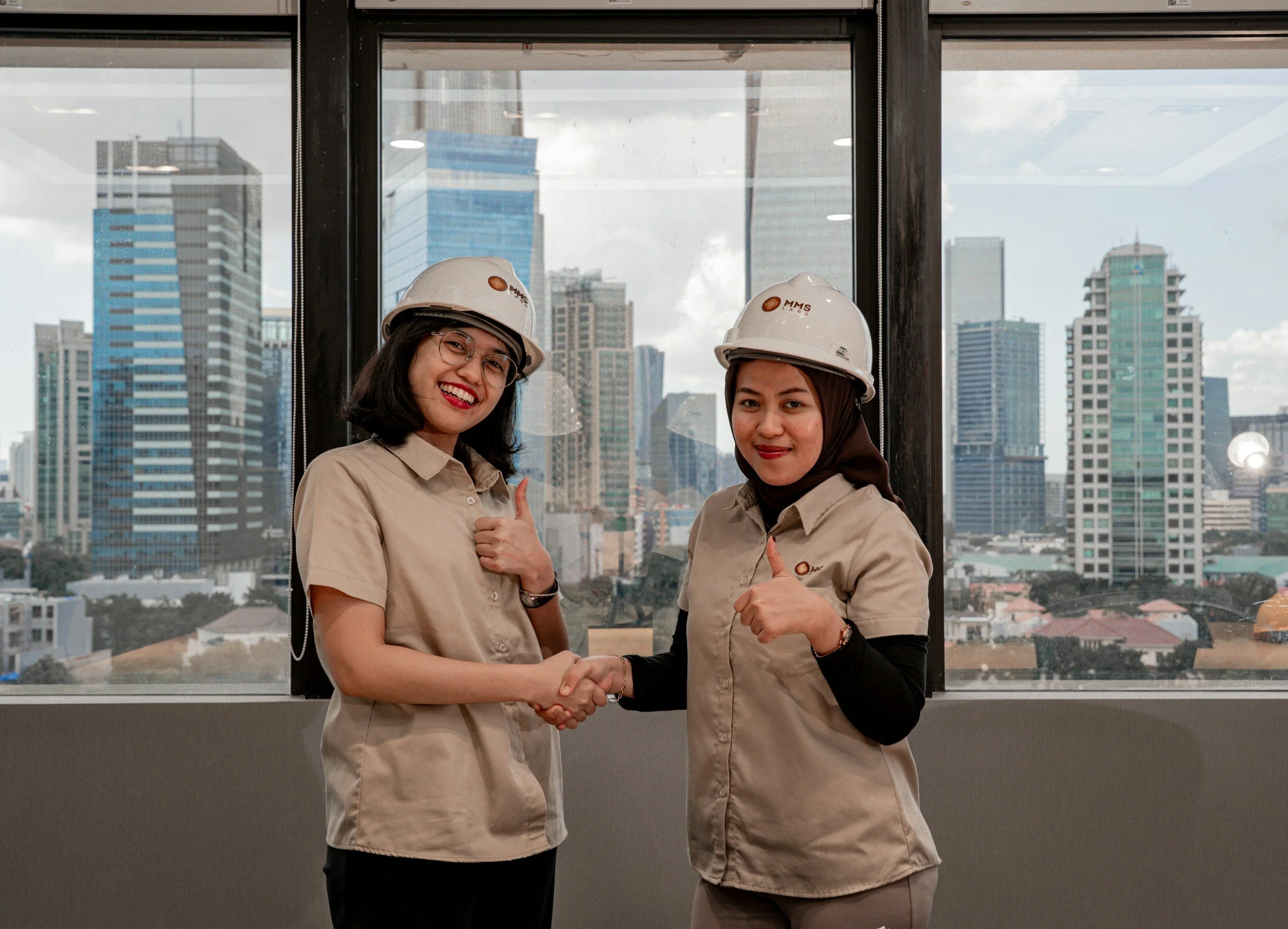 Two women in beige shirts and white safety helmets shaking hands in front of a window with a cityscape of tall buildings.