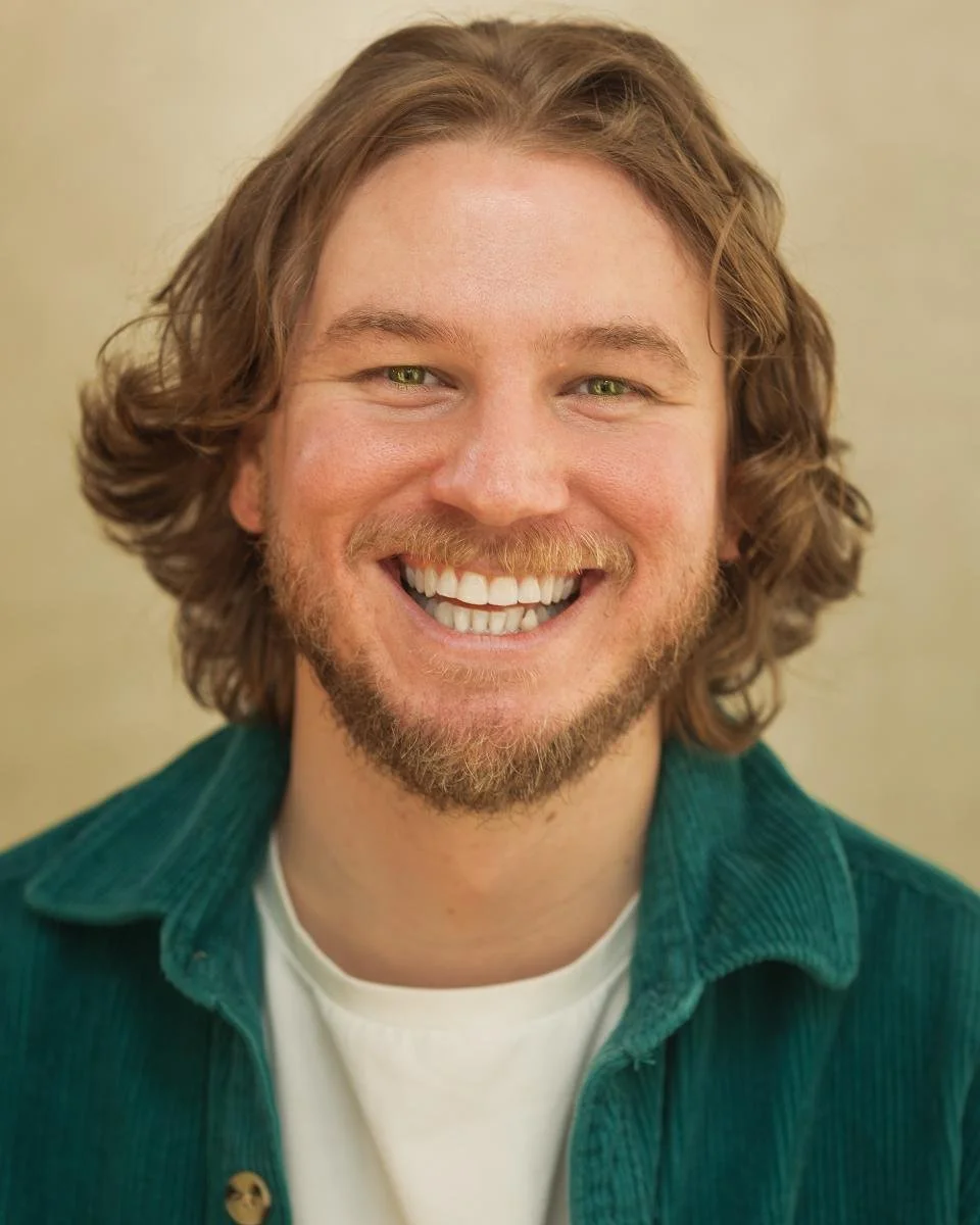 Travis Holmes who is an Actor.  A headshot of a smiling man with curly, shoulder-length brown hair and a beard, wearing a green jacket over a white t-shirt.