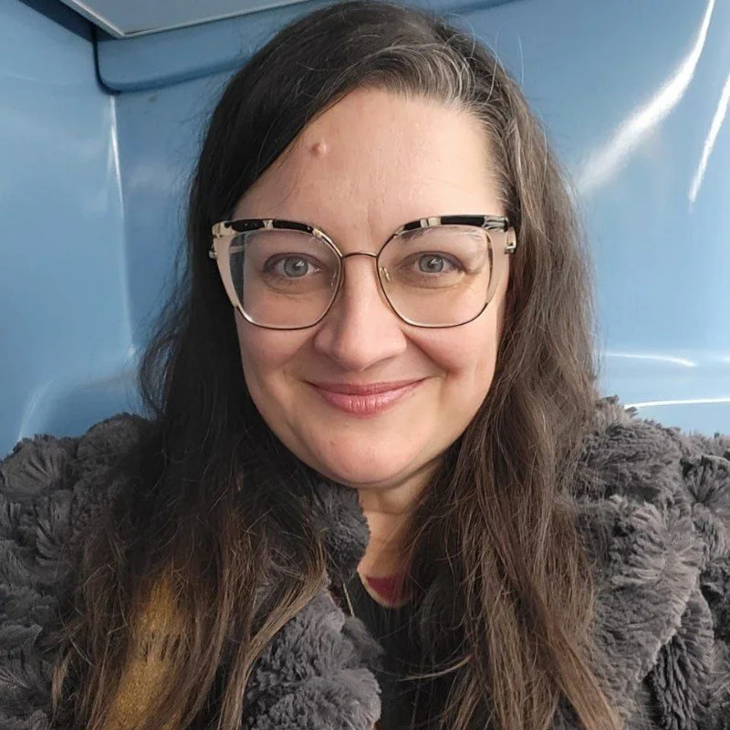 Skye Crawford who is a woman with long brown hair and glasses smiling at the camera, wearing a dark furry coat, indoors with blue walls.