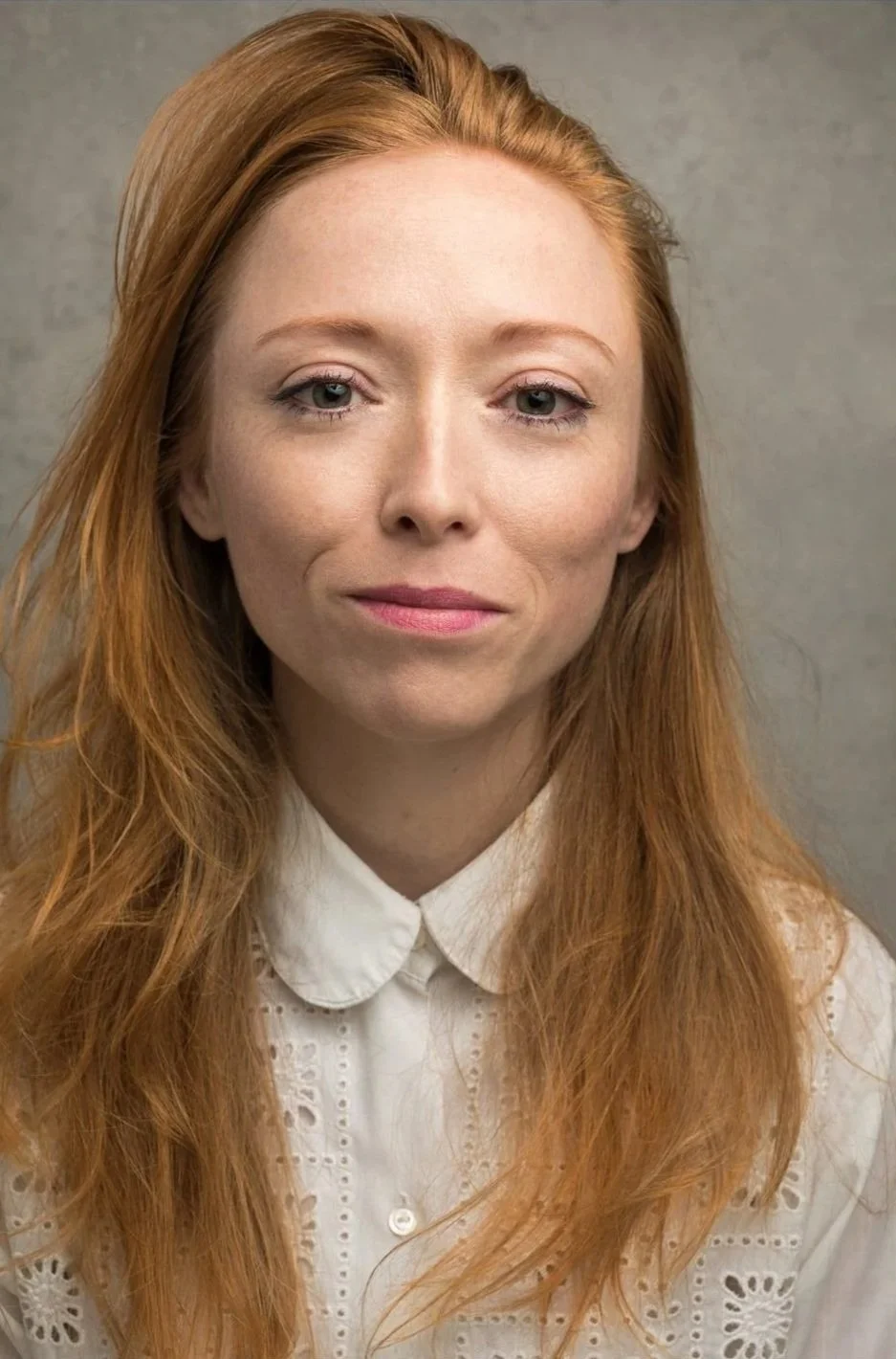 Persia Blue who is an Actor. A headshot of a woman with long red hair, wearing a white embroidered blouse, against a gray background.