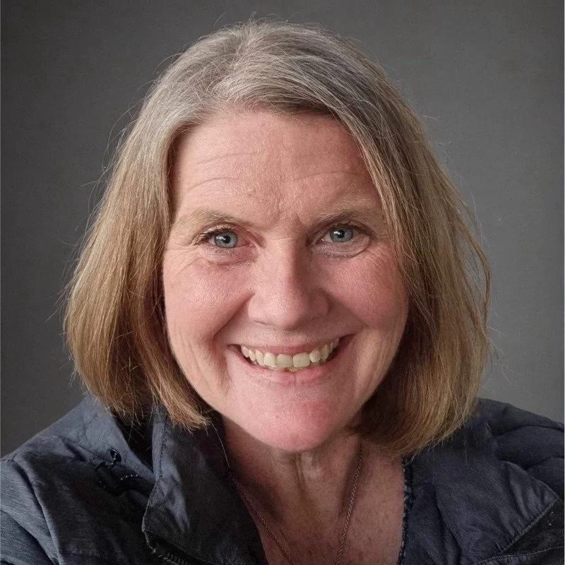 Rachael Presdee. Close-up photo of smiling woman with shoulder-length, light reddish hair, wearing a dark jacket against a gray background.