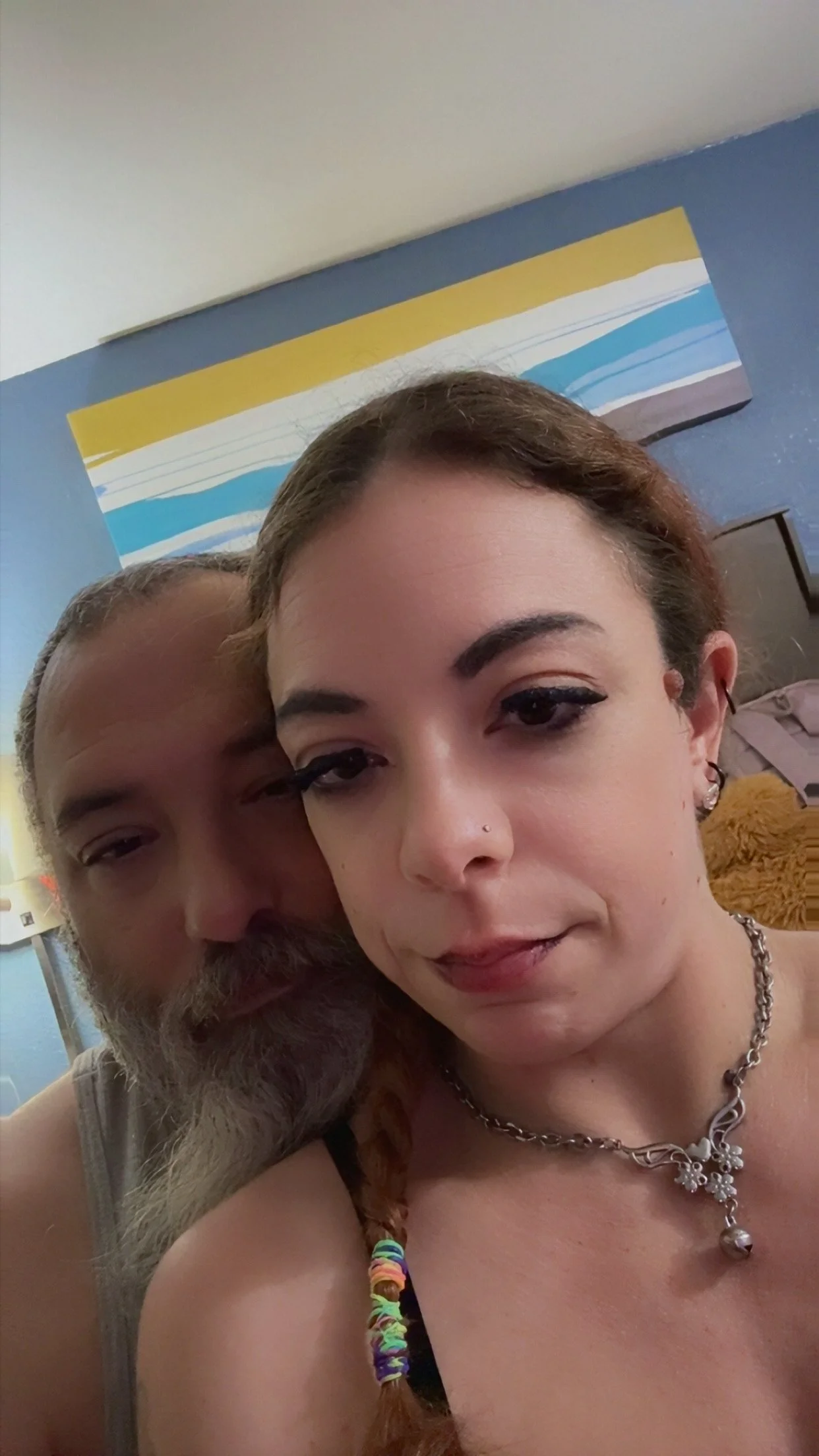 A close-up photo of a woman with short brown hair and tattoos, wearing jewelry and a multicolored beaded bracelet, with a man partially visible next to her. Behind them, there is a blue wall with a colorful, abstract landscape painting.