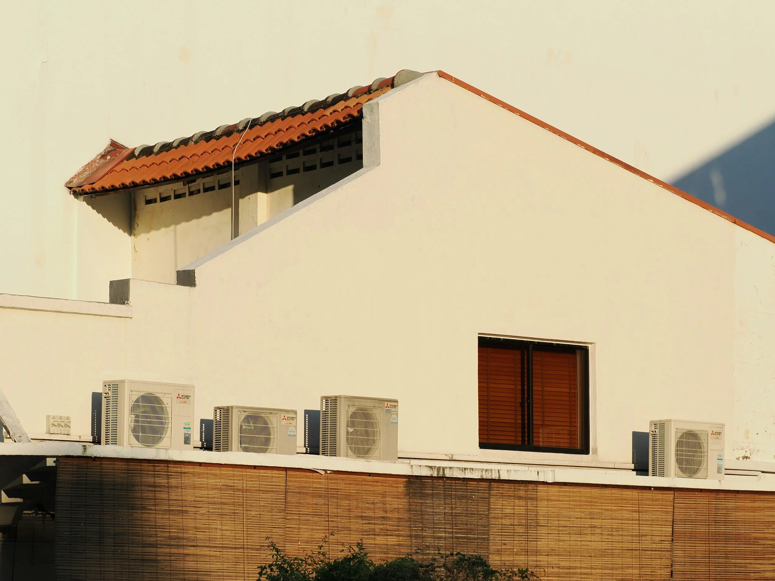 A building with a sloped red-tile roof and multiple air conditioning units mounted on the exterior wall beneath the roofline.