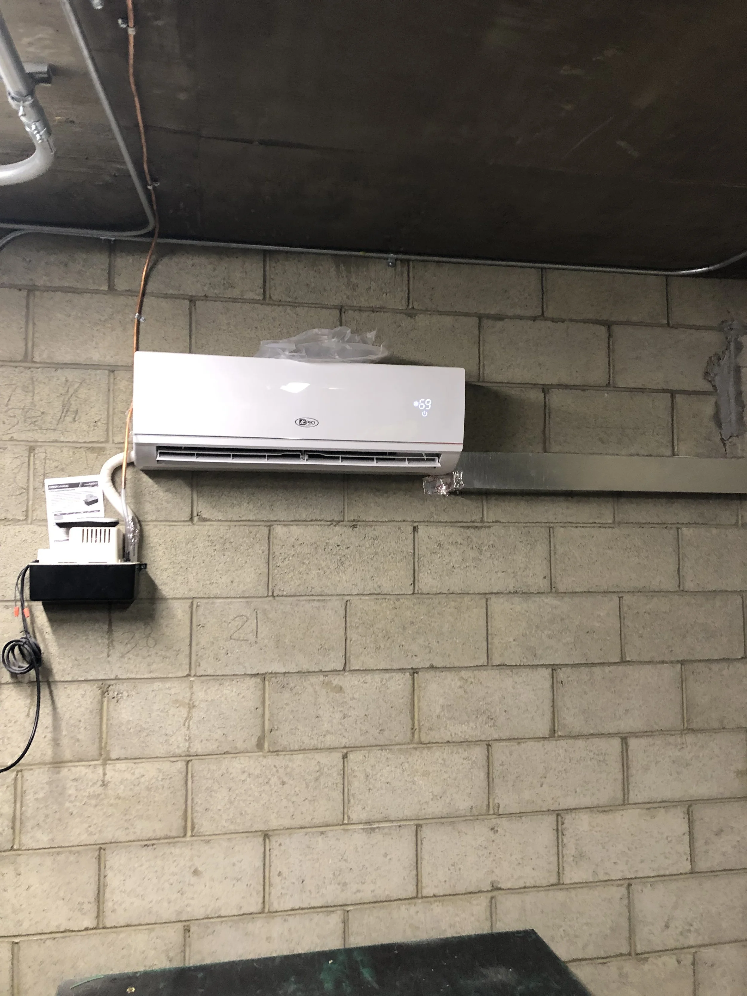 A wall-mounted white air conditioner with a digital display that shows a temperature of 69 degrees, next to electrical wires and devices attached to a concrete block wall.