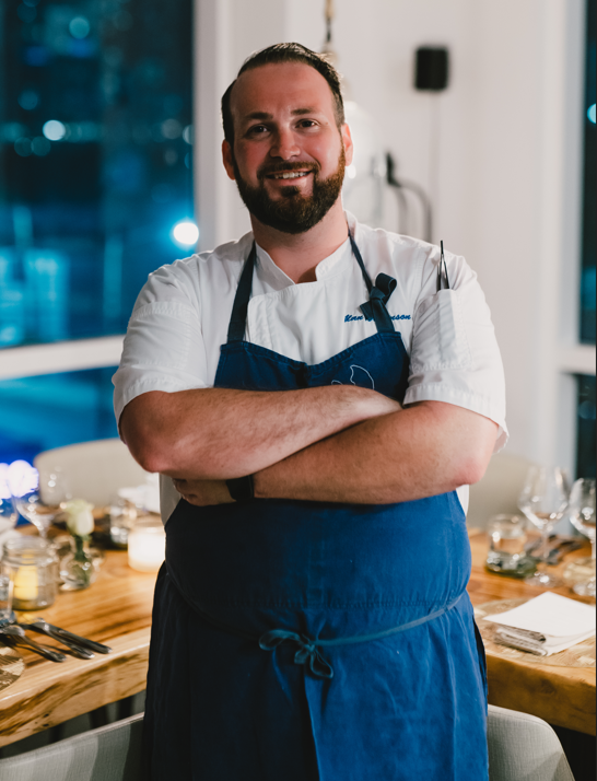 A male chef standing with arms crossed in a restaurant kitchen, wearing a white chef's coat and a blue apron, smiling at the camera.