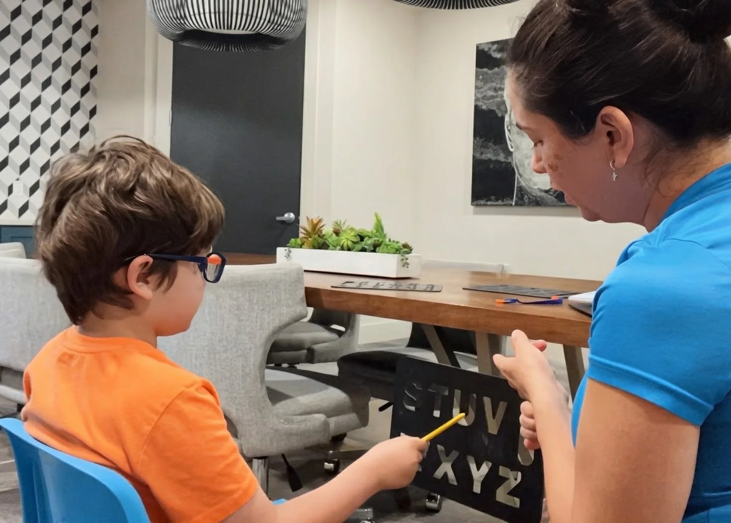 A woman sitting with a young boy at a table. The boy is wearing glasses and an orange shirt, holding a pencil. The woman is wearing a blue shirt. The background includes a table with a succulent plant, some decorative items, and black and white wall art.