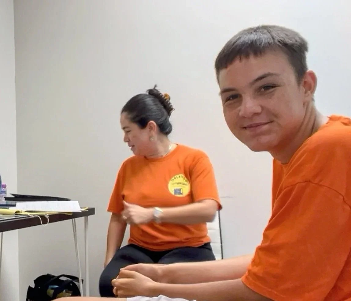 Two people wearing orange shirts sitting in a room with a plain white wall, the woman on the left has her hair in a bun and is looking down, while the young man on the right is smiling at the camera.