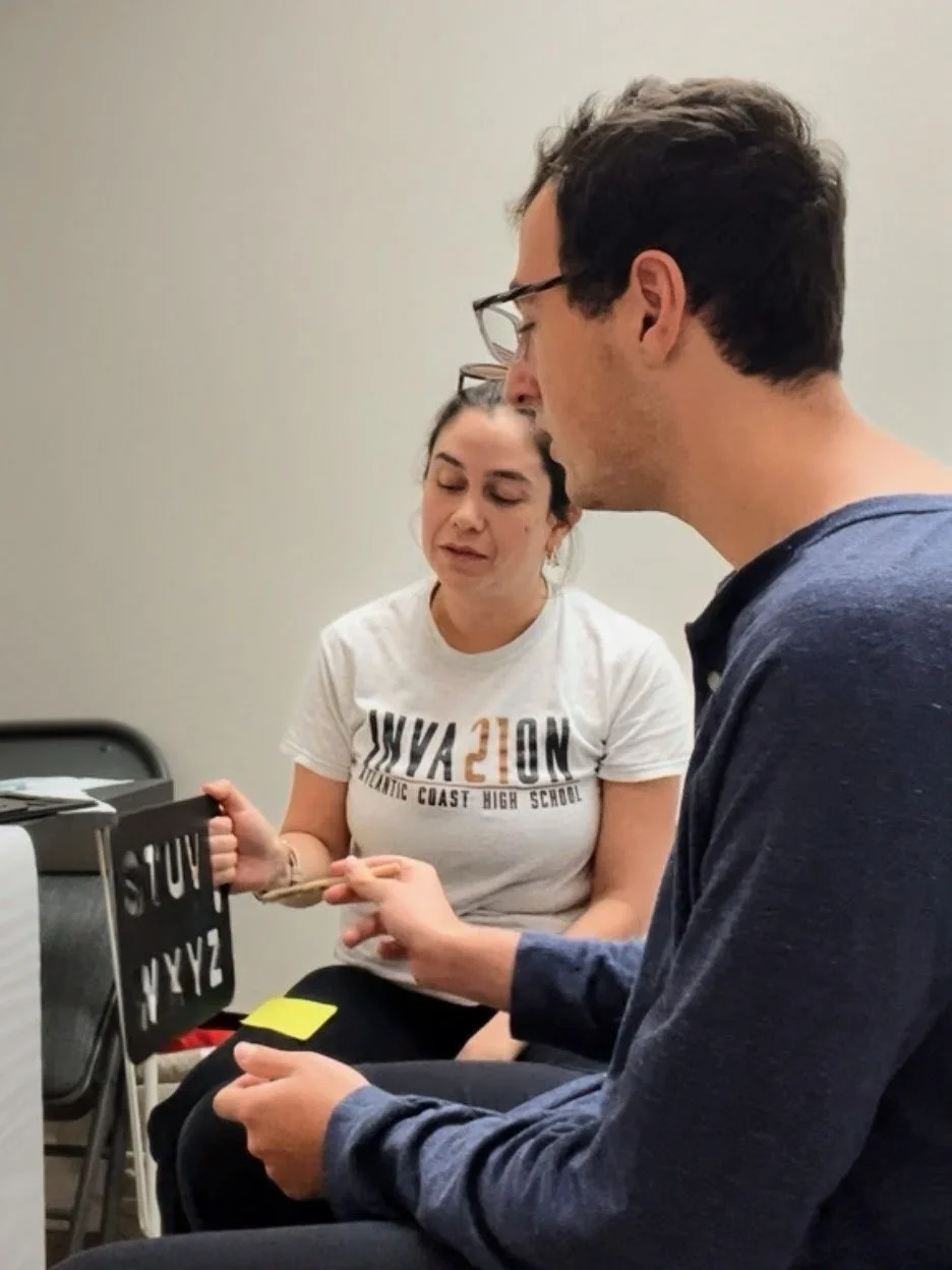 A young woman and young man are sitting. The woman is holding a letterboard, and the man is pointing to the letters with a pencil.