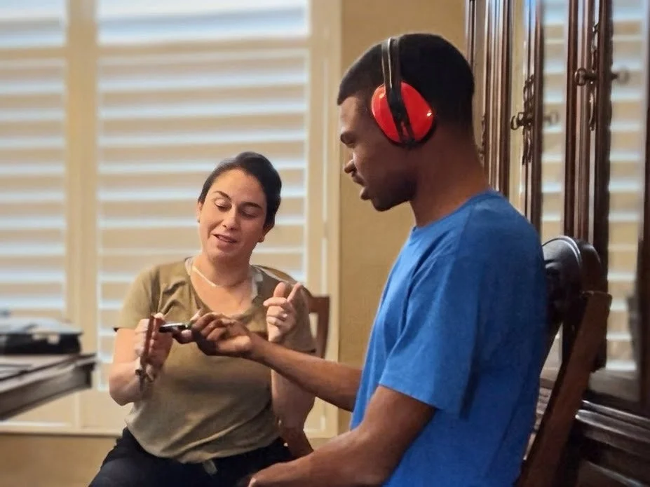 A woman and a man are sitting indoors; the woman is pointing at a phone while the man, wearing large red headphones and a blue t-shirt, is holding the phone and looking at it.