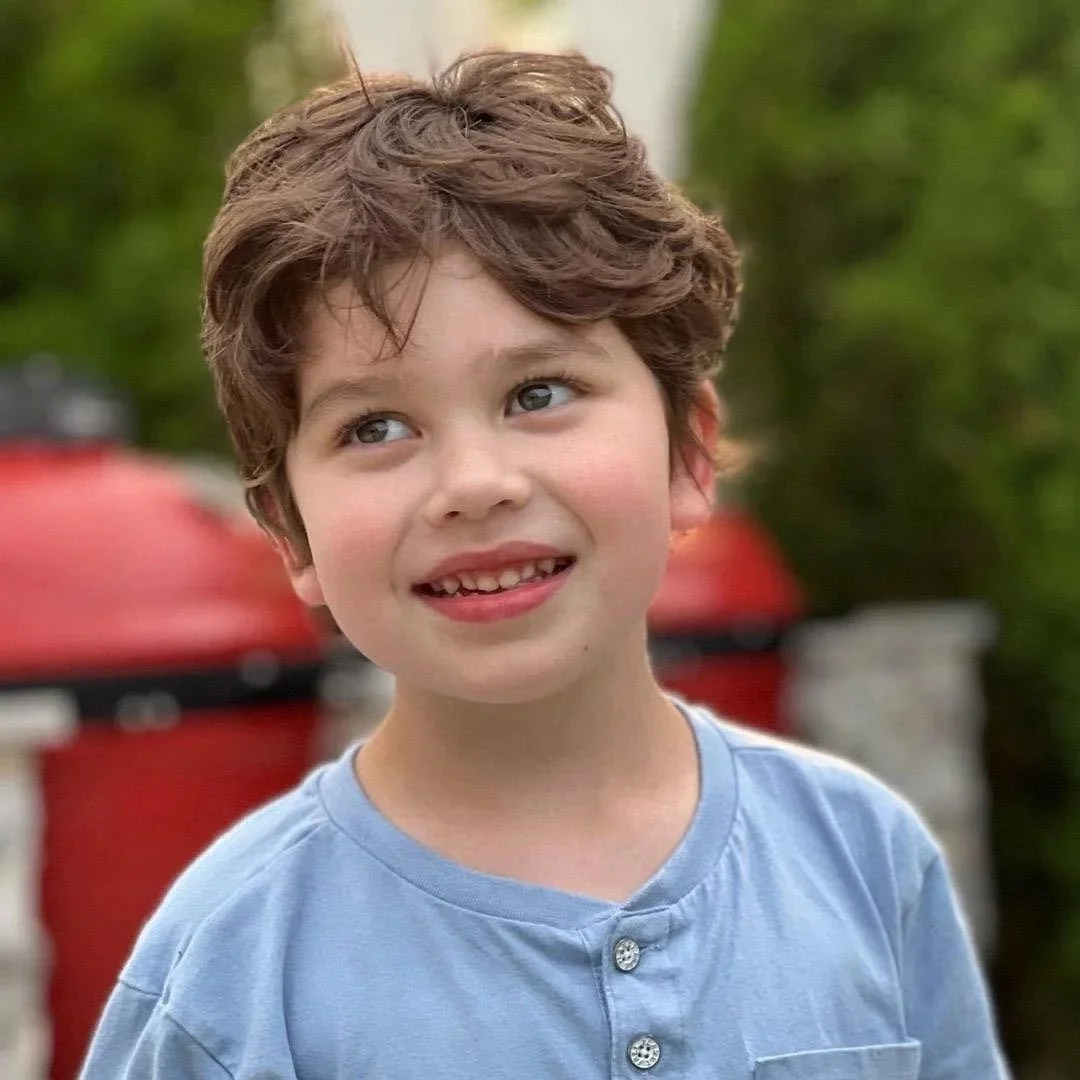 A young boy with curly brown hair, blue eyes, and a missing tooth, smiling outdoors with a red mailbox and green bushes in the background.