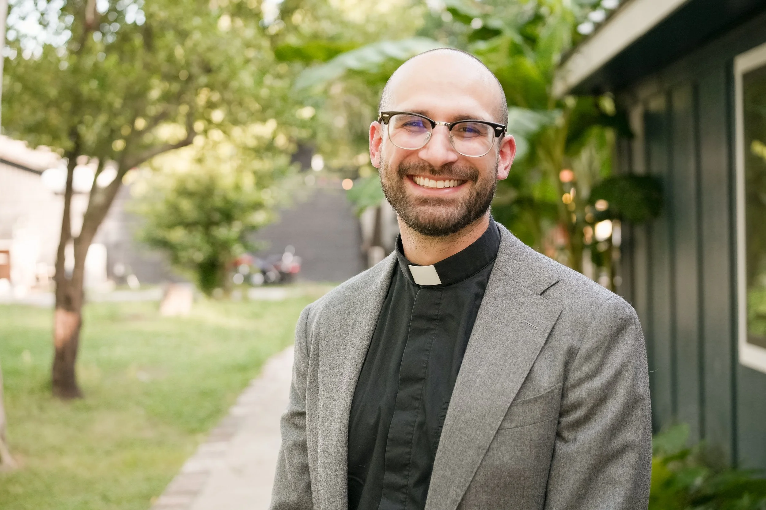 A smiling man with glasses, a beard, and a bald head wearing a clerical collar, a black shirt, and a gray blazer standing outdoors with trees and a house in the background.