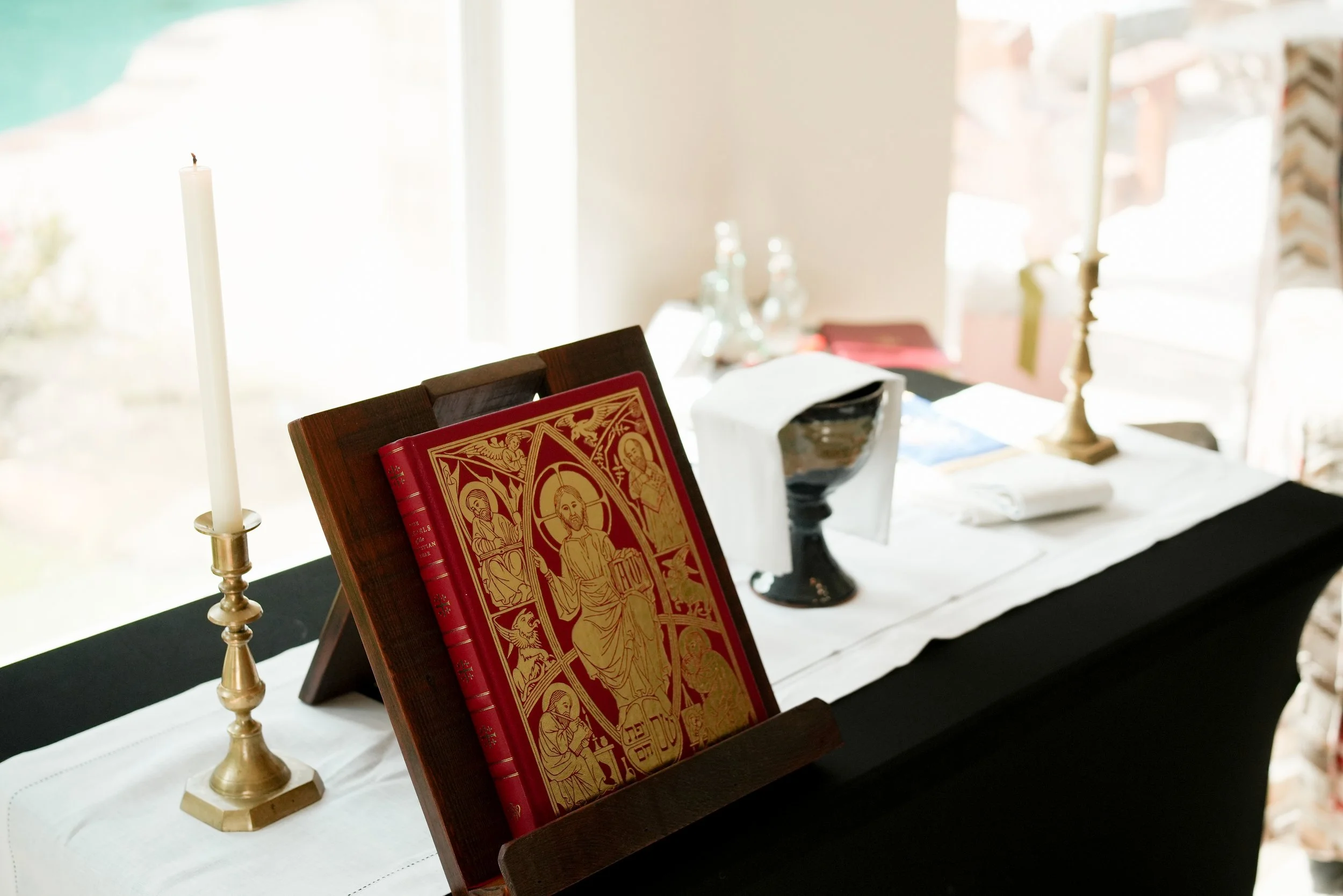 A table with a large red religious book, a tall white candle in a brass holder, and other items set against a window with bright light shining through.