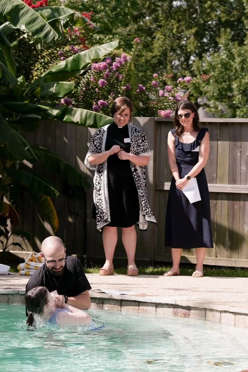 A baptism taking place in a swimming pool outdoors, with a person being baptized by a man, while two women observe from the poolside, one smiling and the other holding papers, with lush green plants and pink flowers in the background.