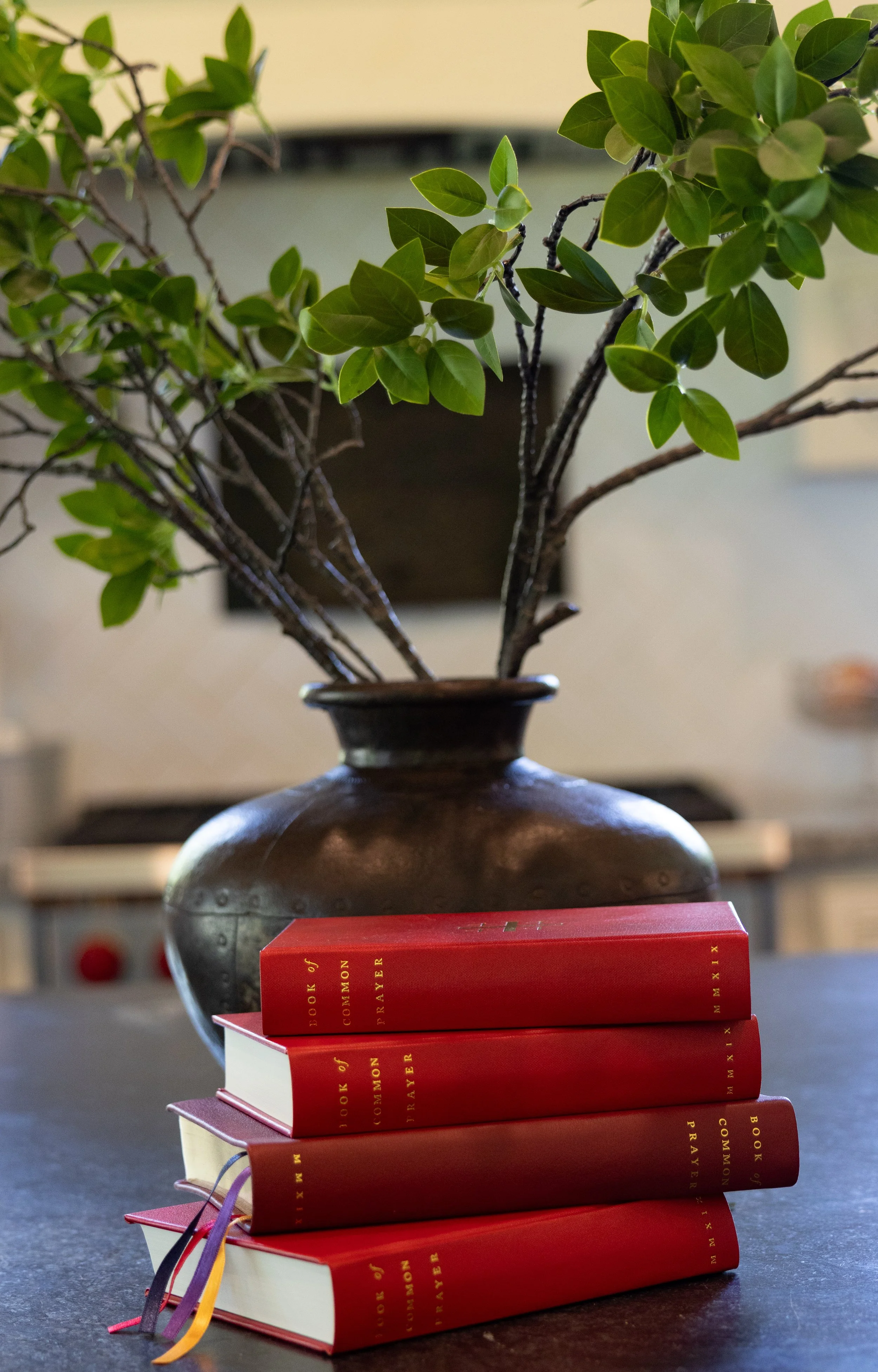 A stack of red Books of Common Prayer with gold lettering on a dark surface in front of a large, brown ceramic vase holding green leafy branches. The background shows a blurred room interior.