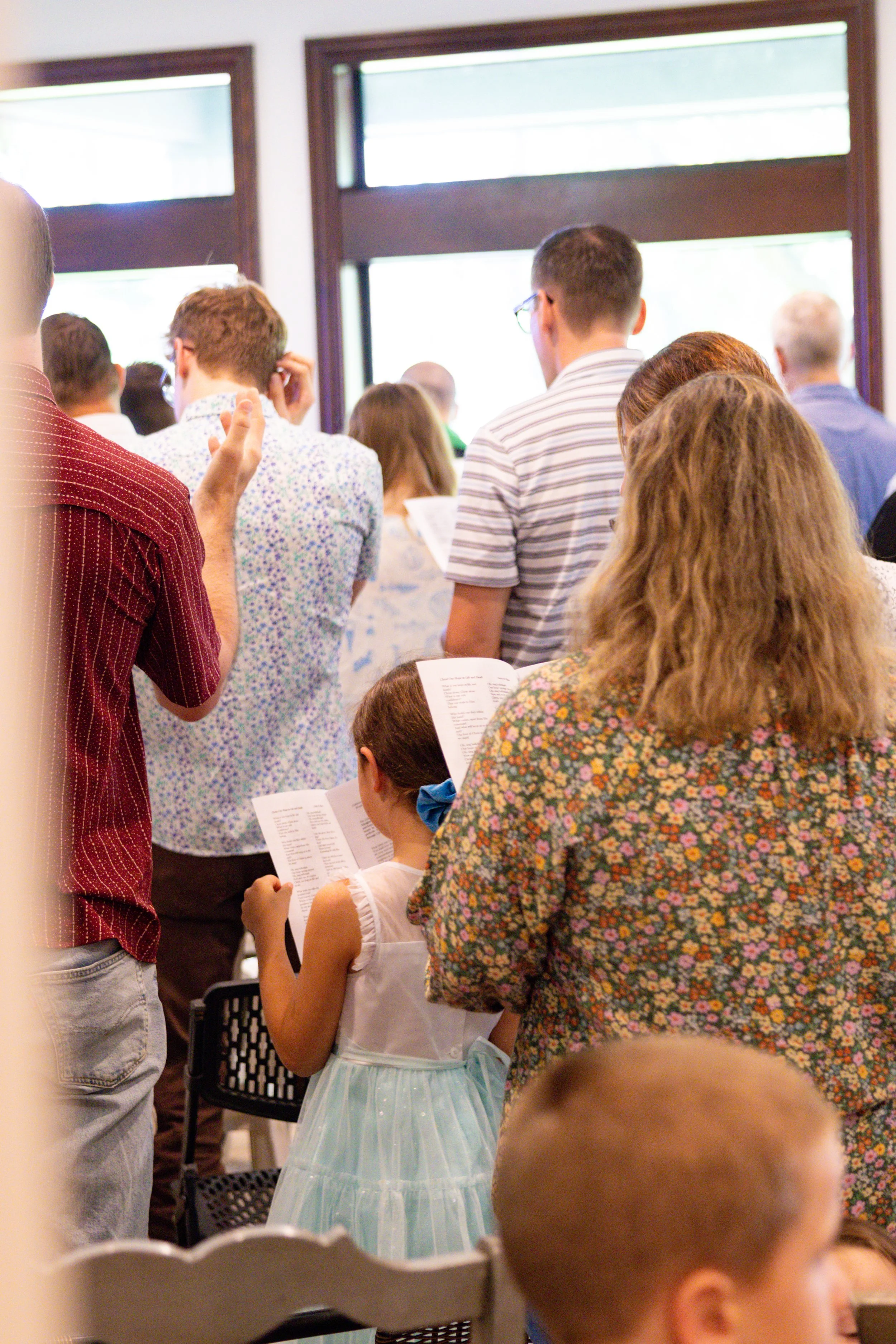 A crowd of people, including children and adults, gathered indoors for worship, with many holding worship bulletins. Large windows in the background let in natural light.