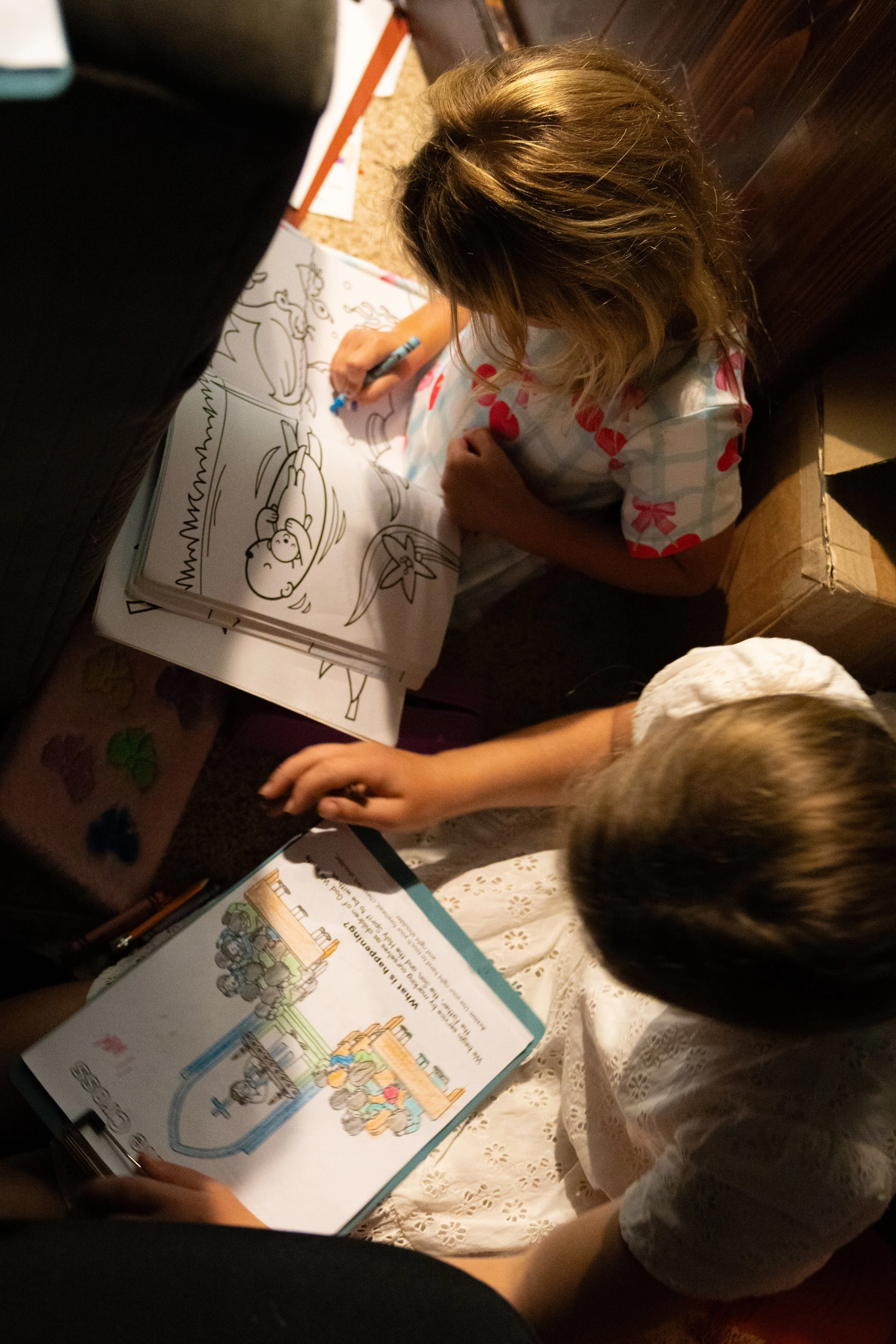 A young girl with brown hair wearing a white dress with red polka dots is sitting on the floor, coloring in a book with a blue marker. There are other books and papers around her.